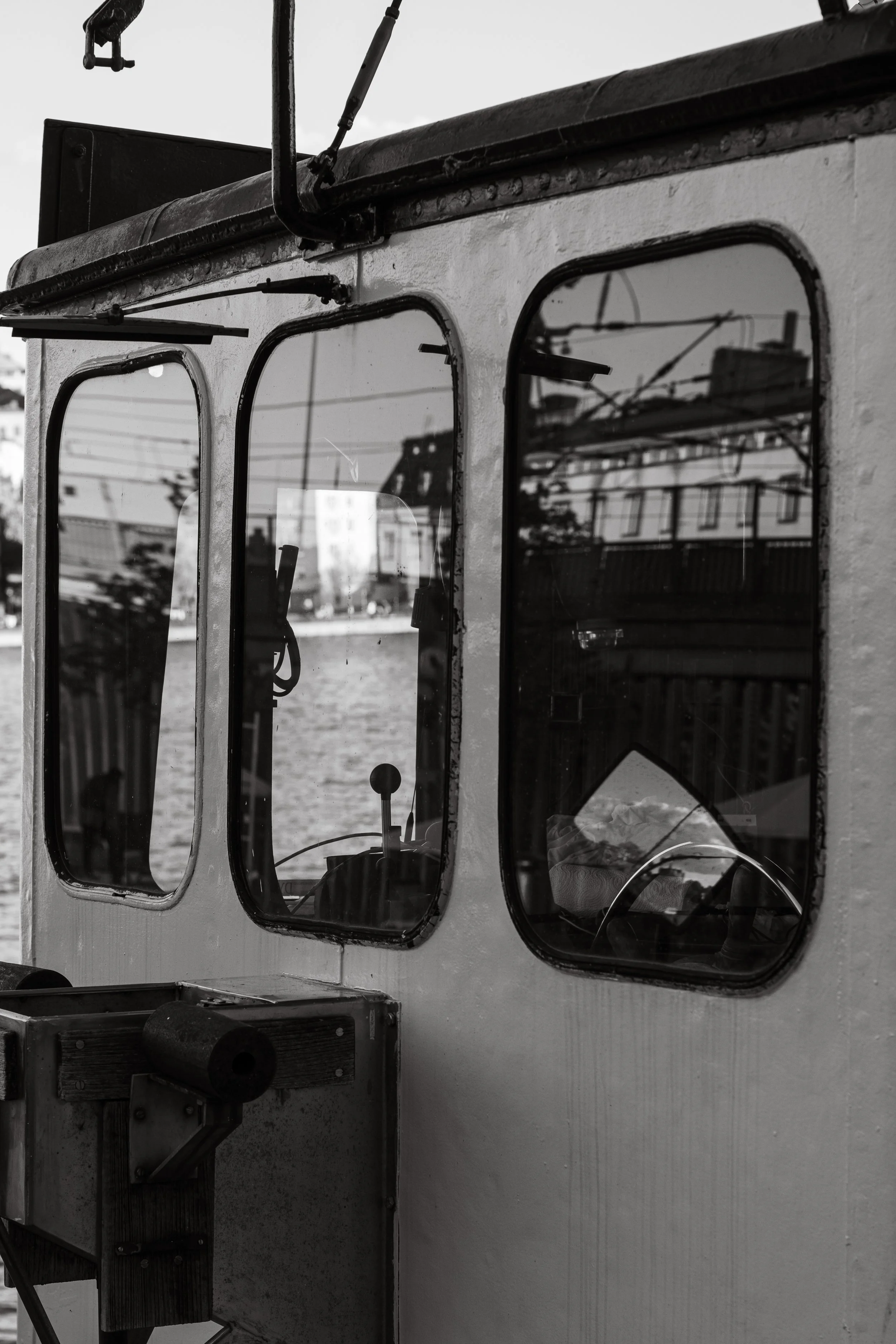 Close-up view of a street vehicle with three rectangular windows, reflections of power lines and buildings visible in the windows, and a mechanical component attached to the exterior. The image is in black and white.
