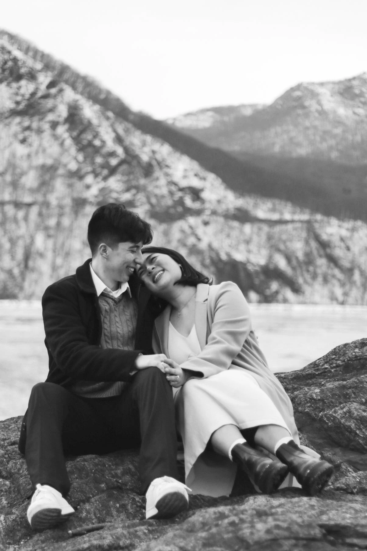 A black-and-white photo of a young couple sitting on rocks outdoors with mountains in the background. They are smiling and leaning into each other, sharing a joyful moment.