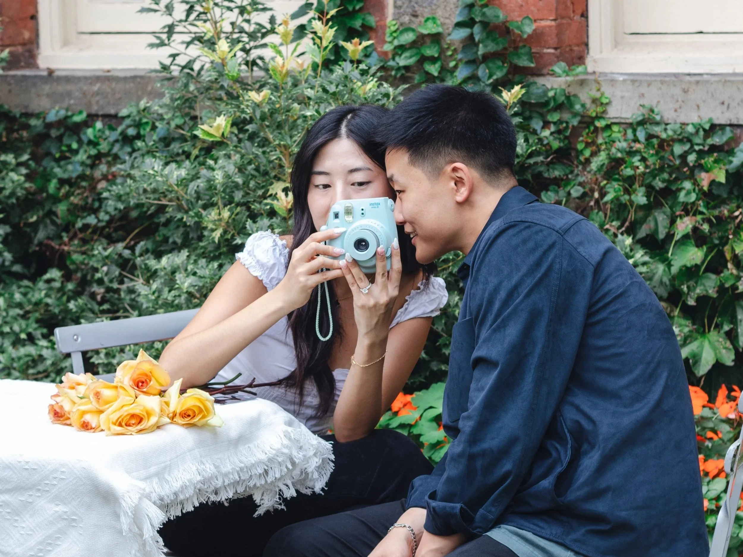 A woman and a man sitting closely together outdoors, looking at a Polaroid camera. There are yellow roses on a table in front of them, with greenery and a brick wall background.