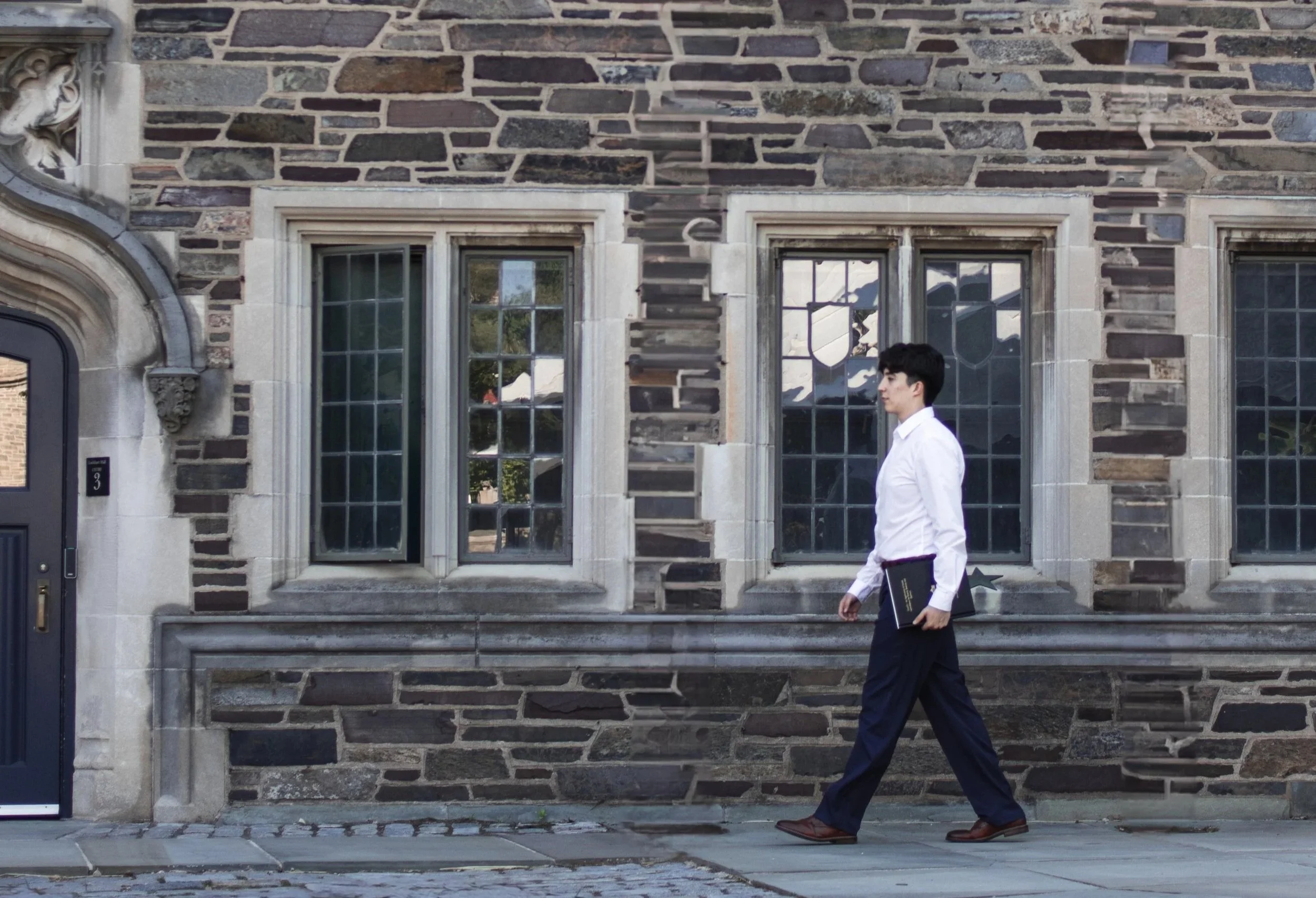 A young man in a white shirt and dark trousers walking past a historic stone building with three windows and an ornate stone archway, holding a folder.