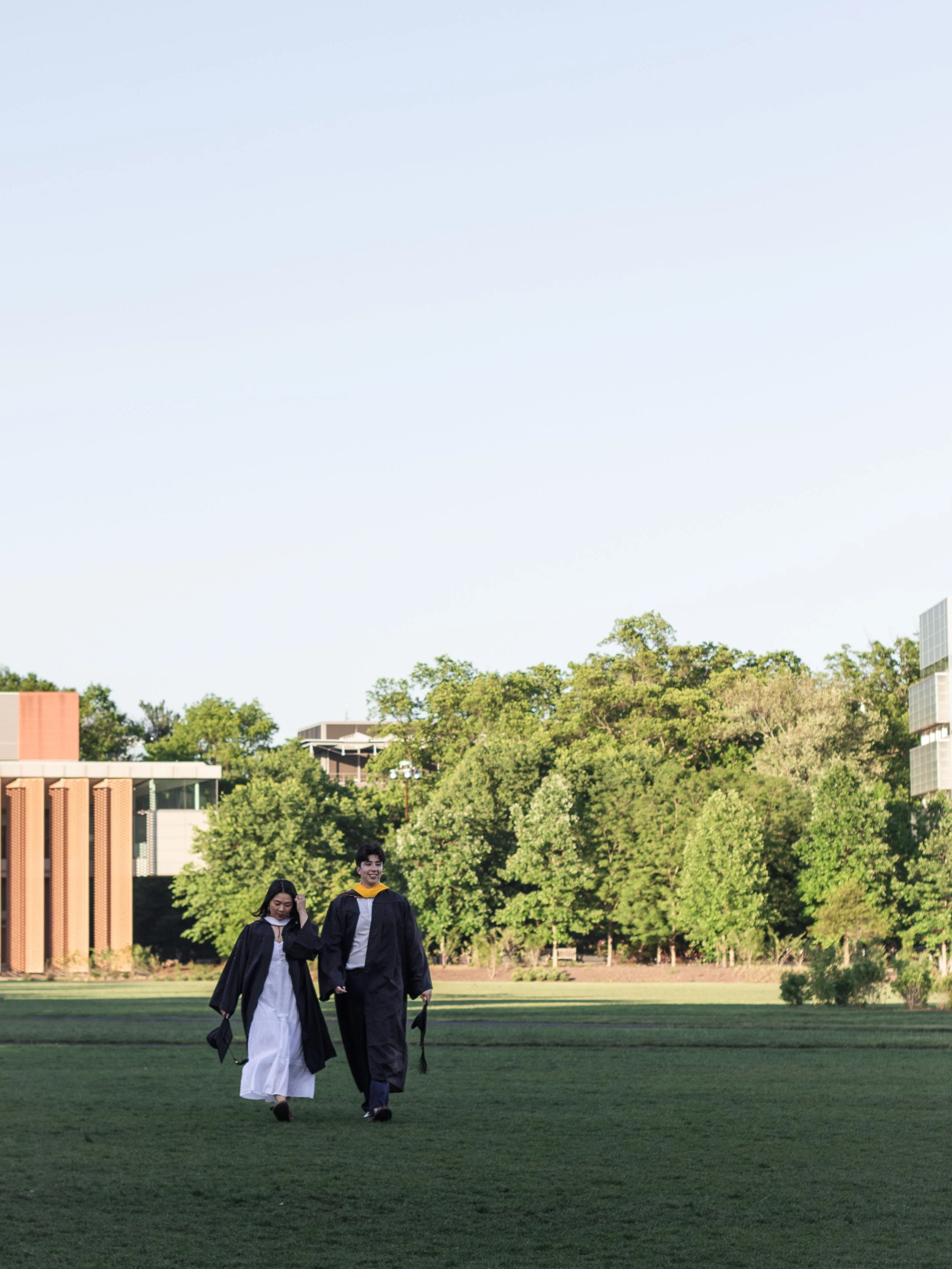 Two graduates in cap and gown walking on a grassy field with trees and buildings in the background, during daytime.