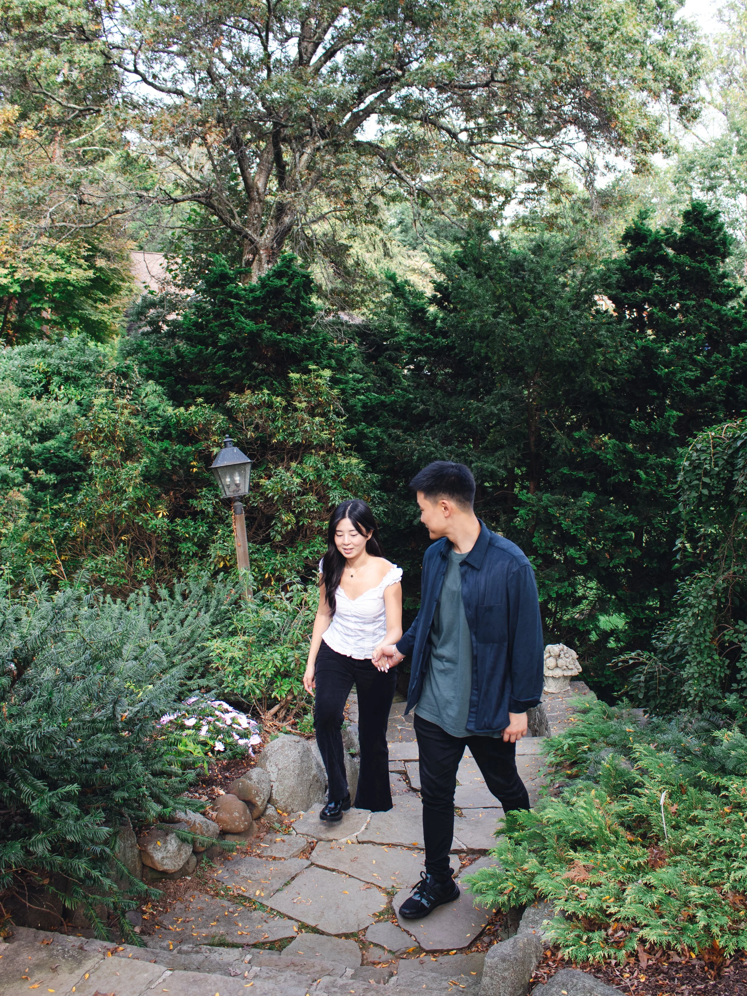 A young man and woman walking hand in hand along a stone pathway in a lush, green garden surrounded by trees and plants. The woman has long dark hair and is wearing a white top with black pants, while the man has short dark hair and is dressed in a dark jacket and pants. A small lamp post is visible on the left side of the path.
