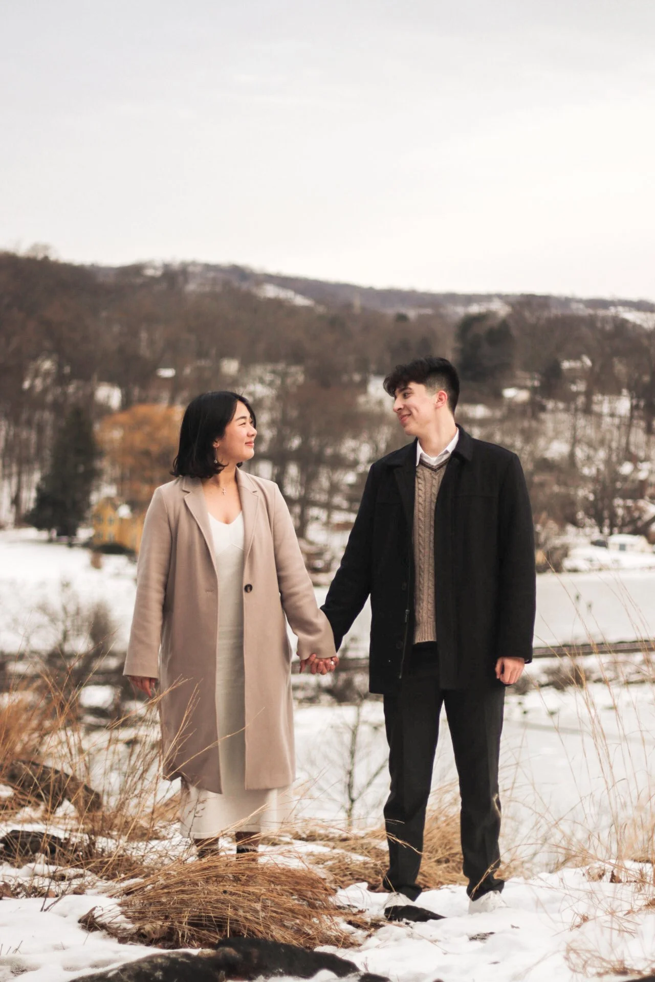 A couple holding hands and smiling at each other while walking on snow-covered ground outdoors in winter, with trees and hills in the background.