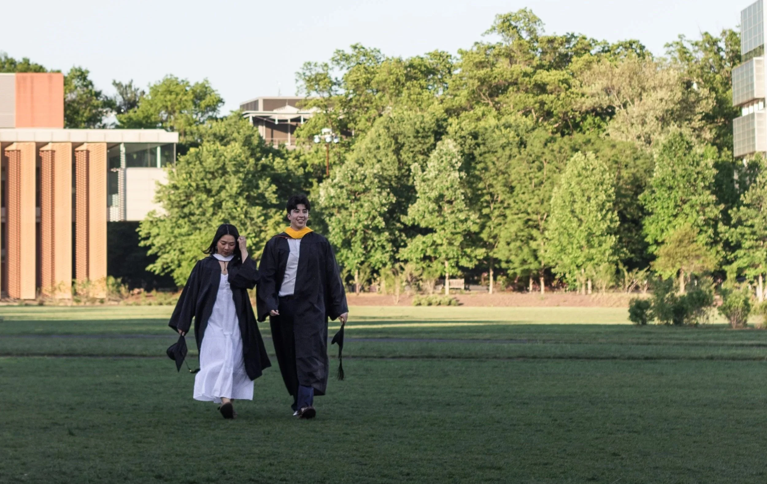 Two graduates walking across a grassy campus field with trees and modern buildings in the background, one male and one female, both wearing black caps and gowns, smiling and holding hands.