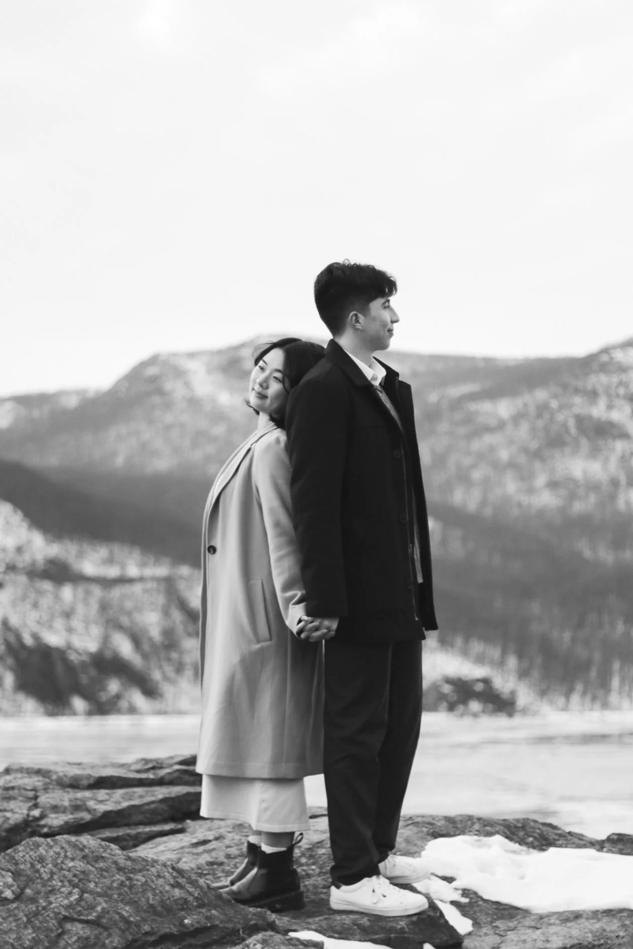 A black and white photo of a young couple standing outdoors on rocks with mountains in the background. The woman is leaning her head on the man's shoulder, both holding hands, looking in opposite directions.