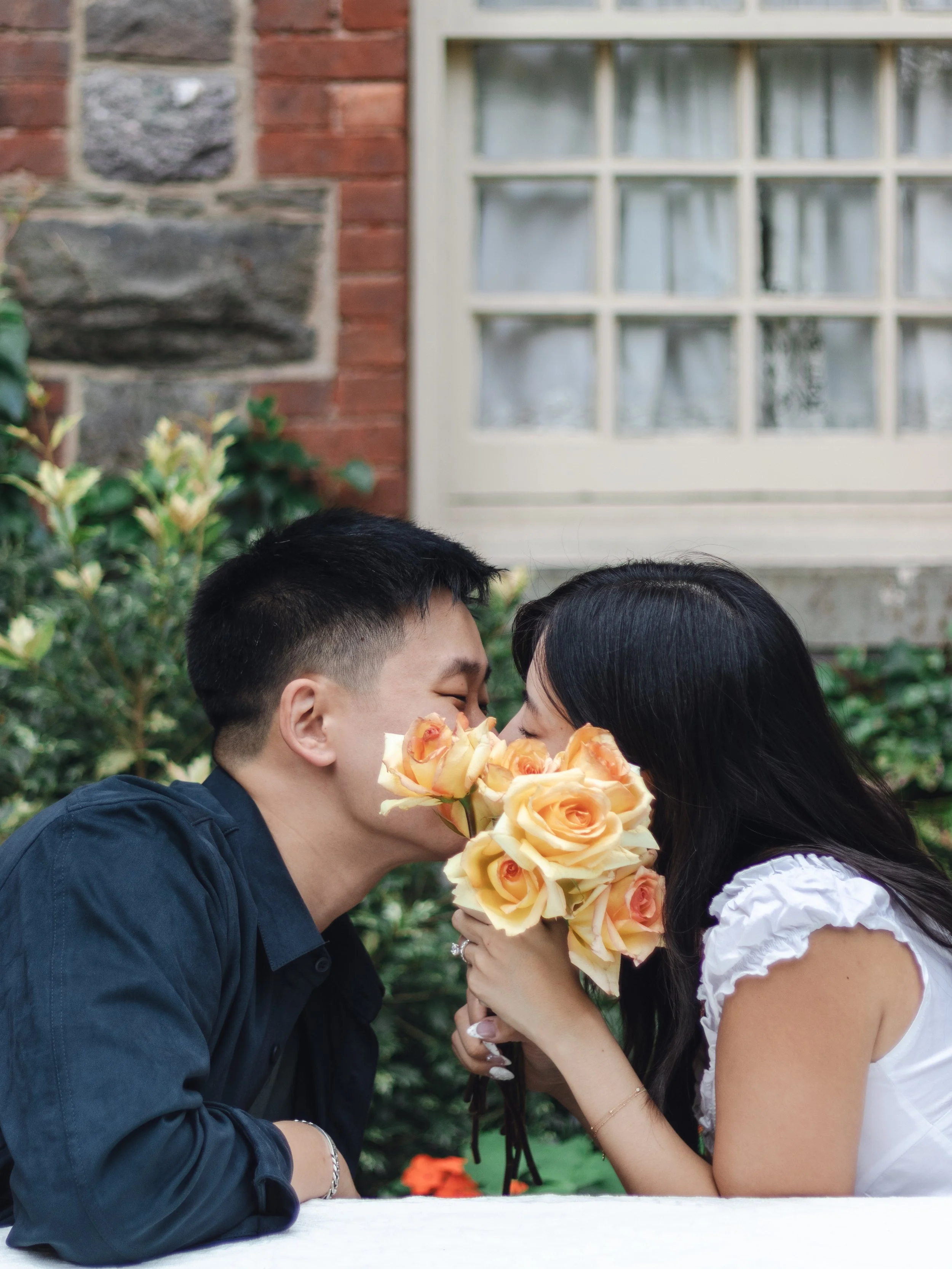 A couple appears to be sharing a romantic moment, with the woman holding a bouquet of yellow roses and both about to kiss outdoors.