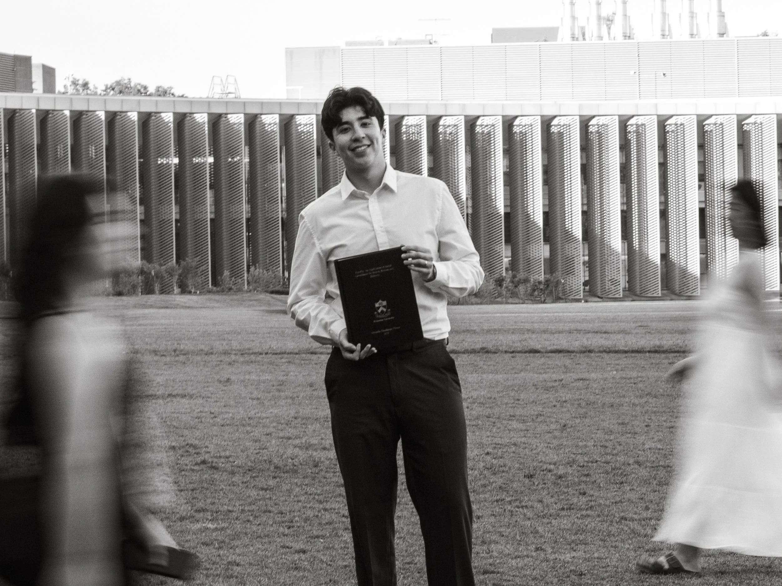 A young man in a white shirt holding a book standing outdoors with modern building in the background.