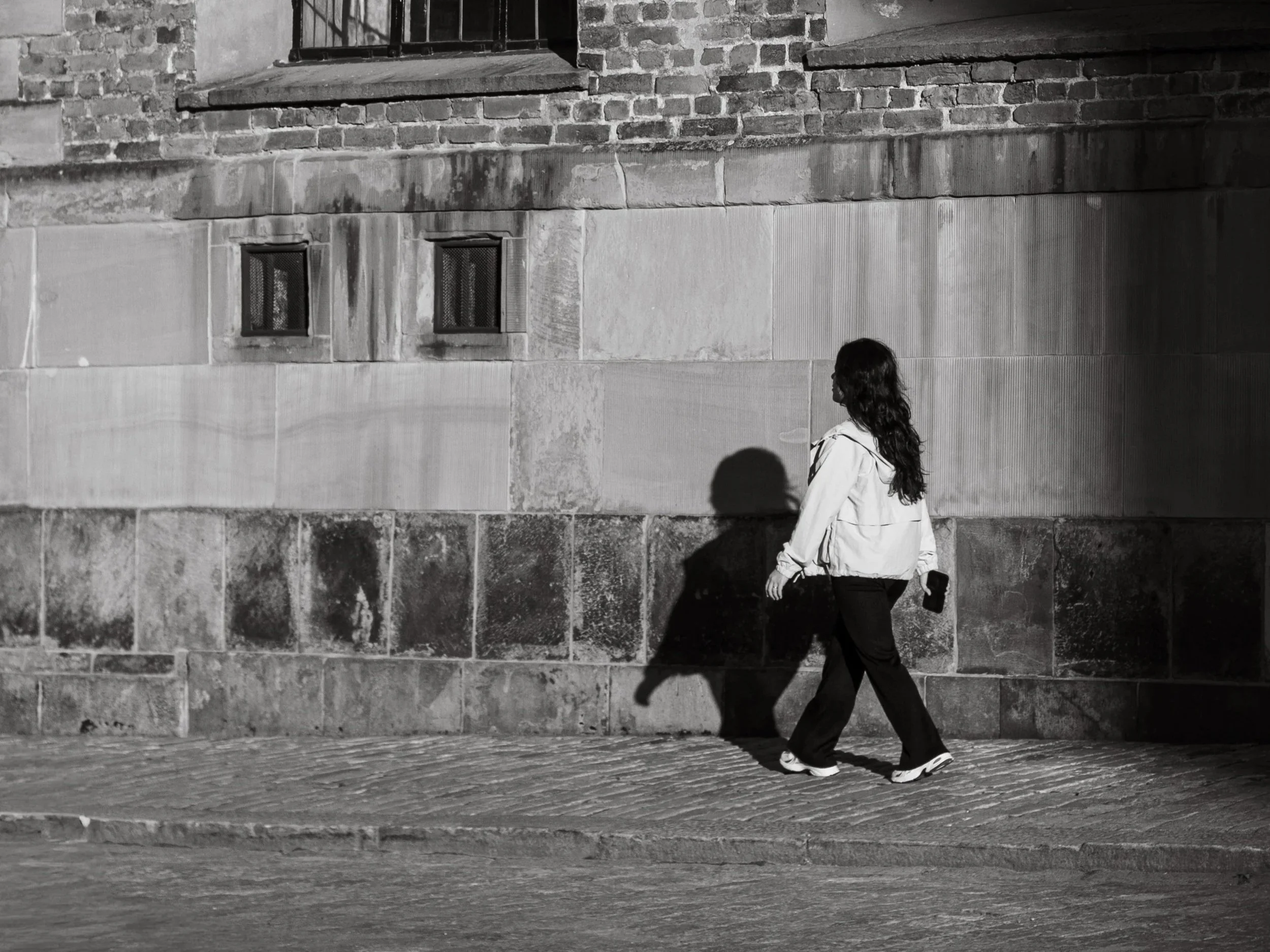 A woman walking on a city sidewalk at night next to a brick building with small windows, cast in shadows with her hair flowing and holding a phone in her right hand.