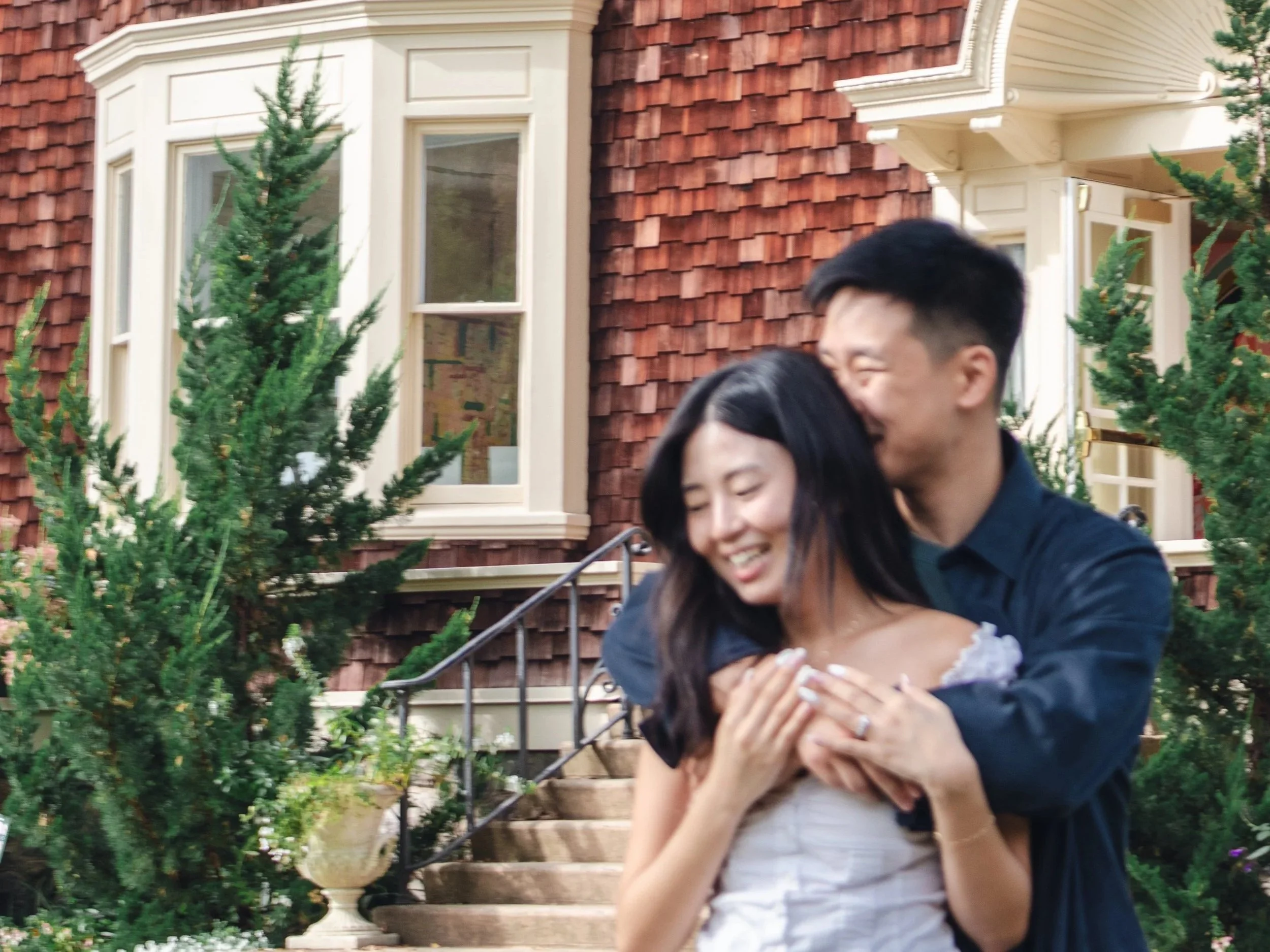 A couple hugging and smiling outdoors in front of a house with wooden shingles and large windows, surrounded by greenery and trees.