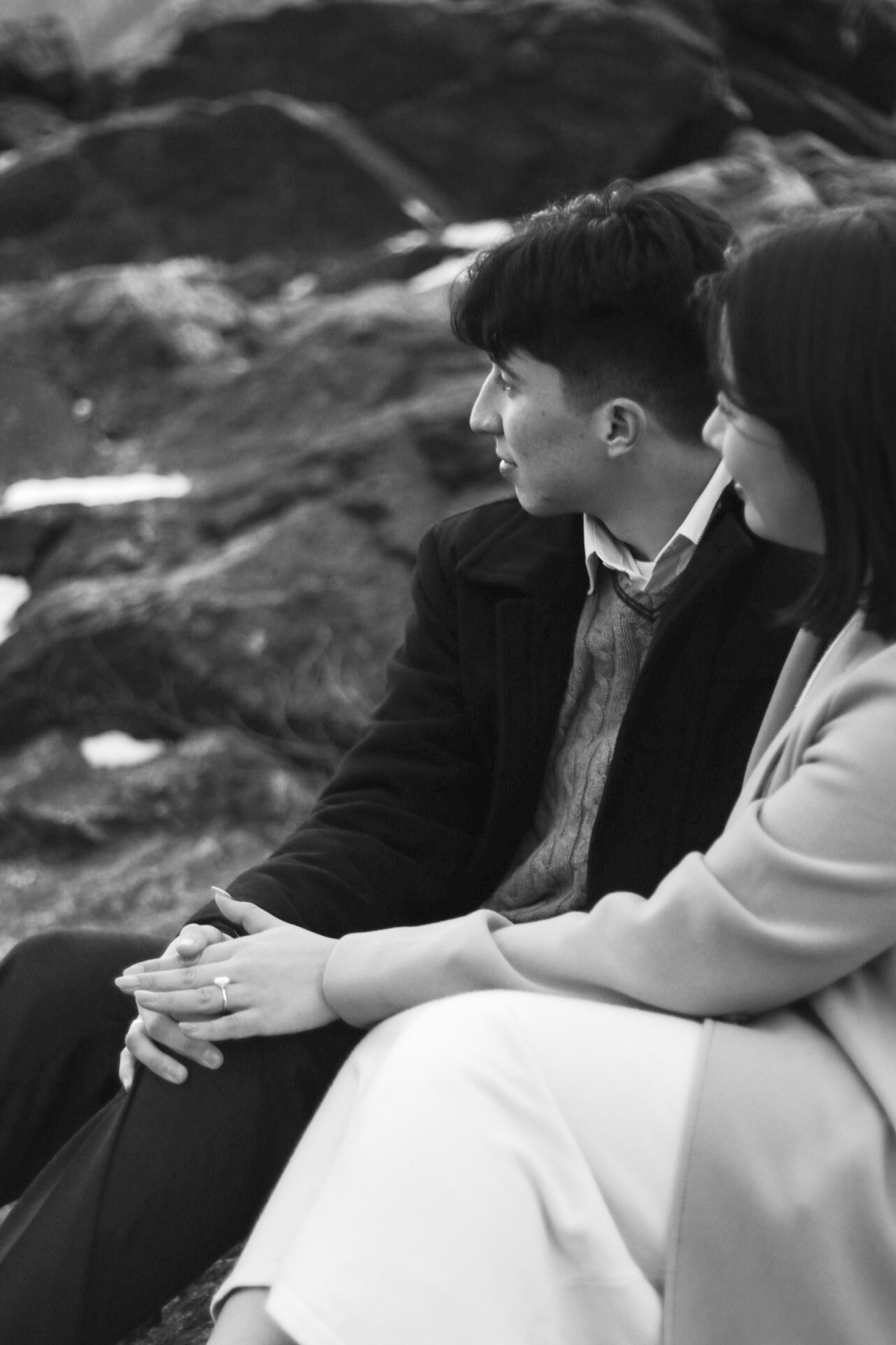 Black and white photo of a man and woman sitting outdoors on a rocky surface, with their hands clasped and looking to the side.