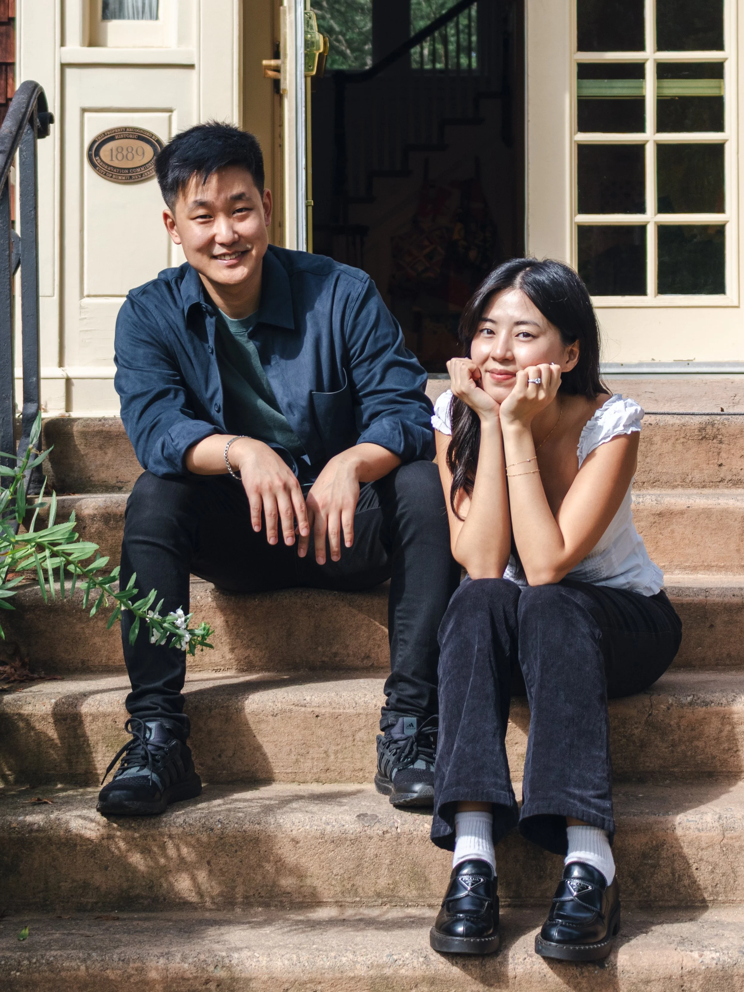 A young man and woman sitting on the steps outside a house, smiling at the camera. The man is wearing a dark blue jacket and black pants, and the woman is wearing a white top and black pants. The house has cream-colored walls and a door with a window. There are plants around the steps.
