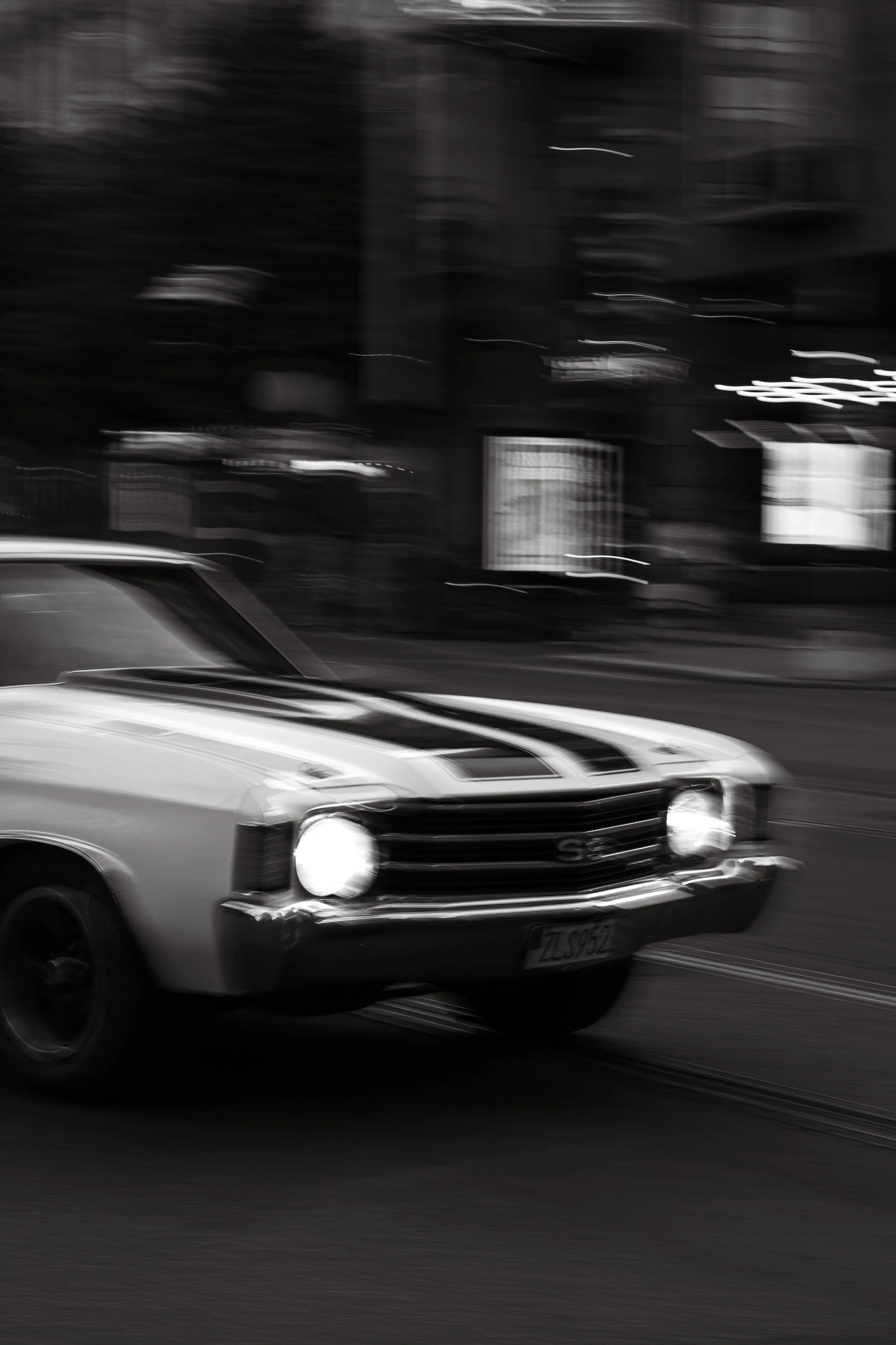 A black and white photograph of a classic car driving on the street, with a blurred cityscape in the background.