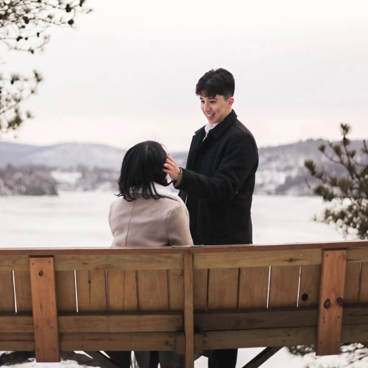 A young man tenderly touches a woman's face in an outdoor winter setting, with a wooden fence, snow, water, and distant hills in the background.