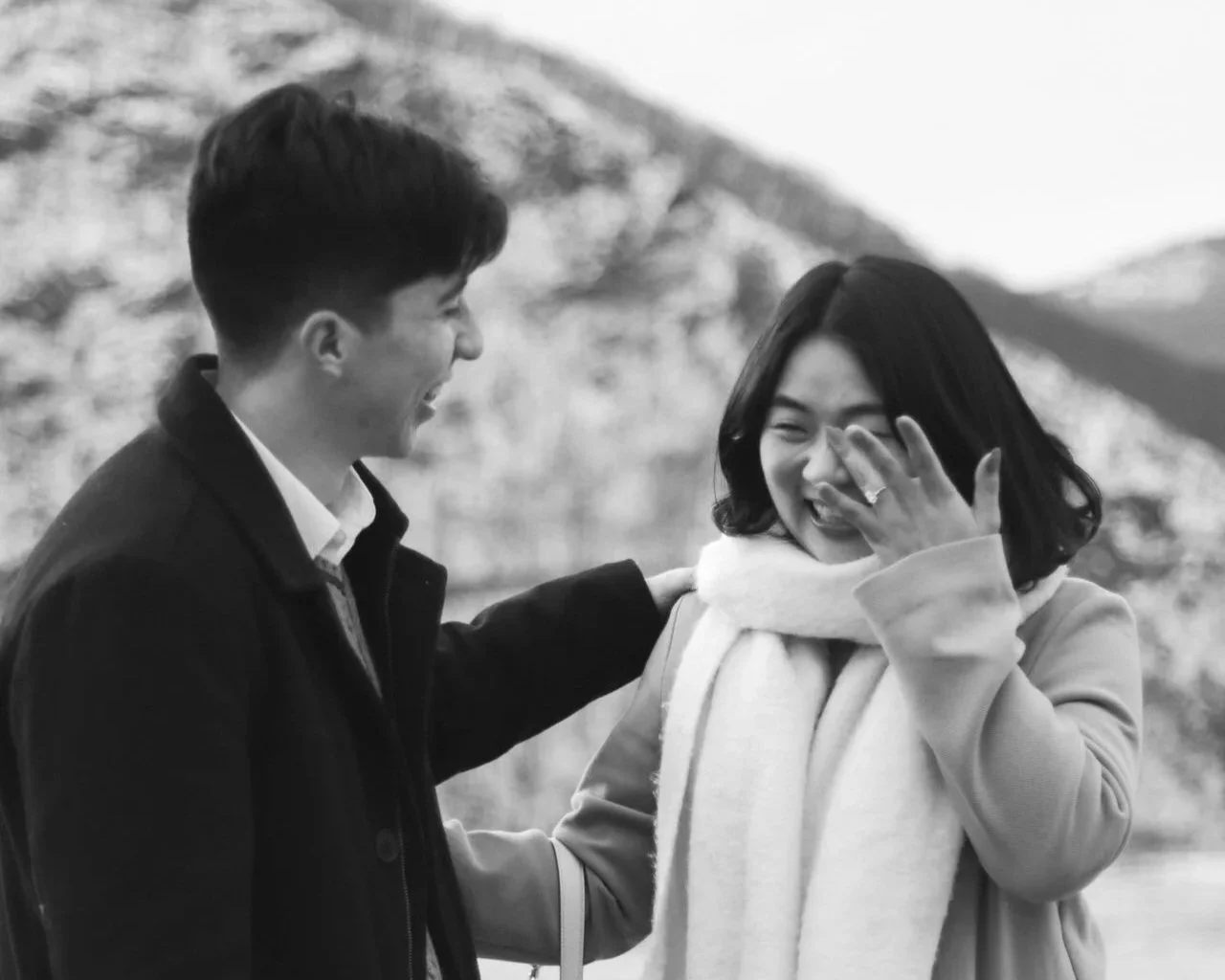 A black and white photo of a man and woman smiling and sharing a joyful moment outdoors, with mountains in the background.