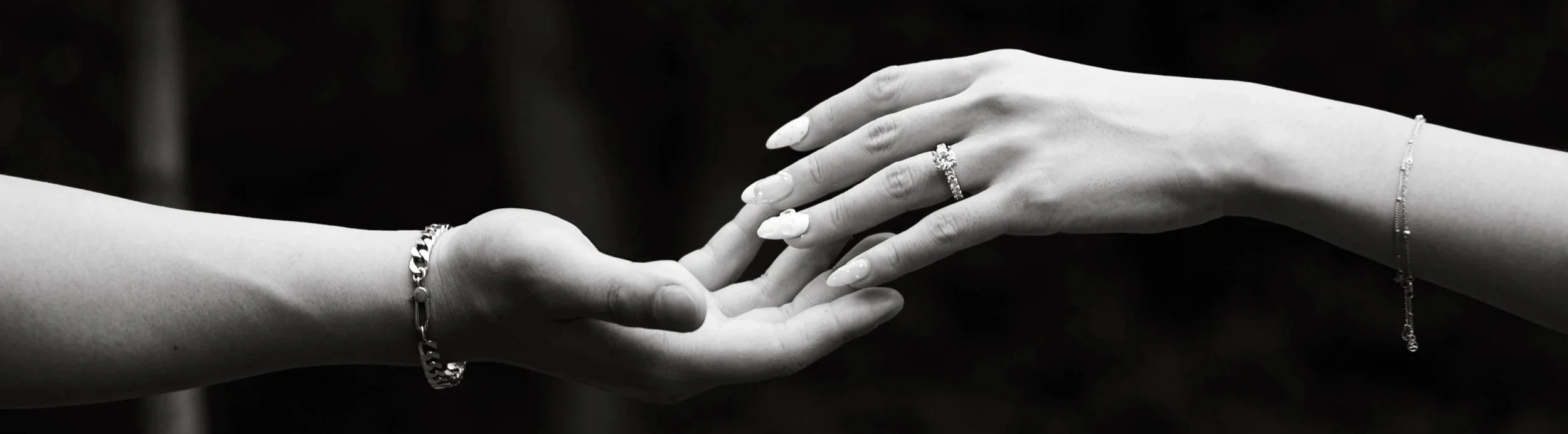 Two hands reaching towards each other, with one hand wearing a ring and bracelet, the other wearing a bracelet, in black and white.