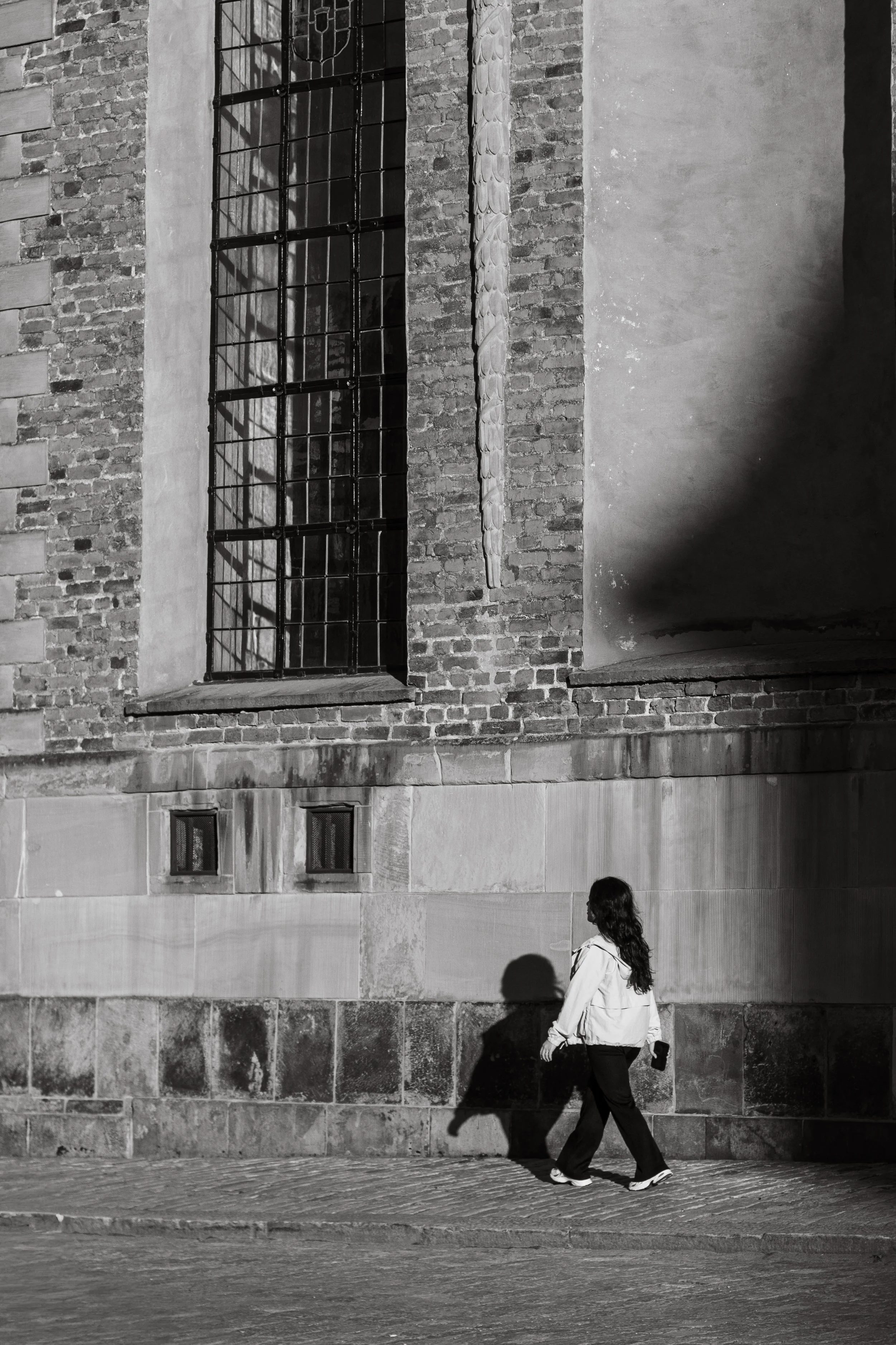 A woman with long hair walking along a city sidewalk at night, with a shadow cast on a brick wall from nearby streetlights.