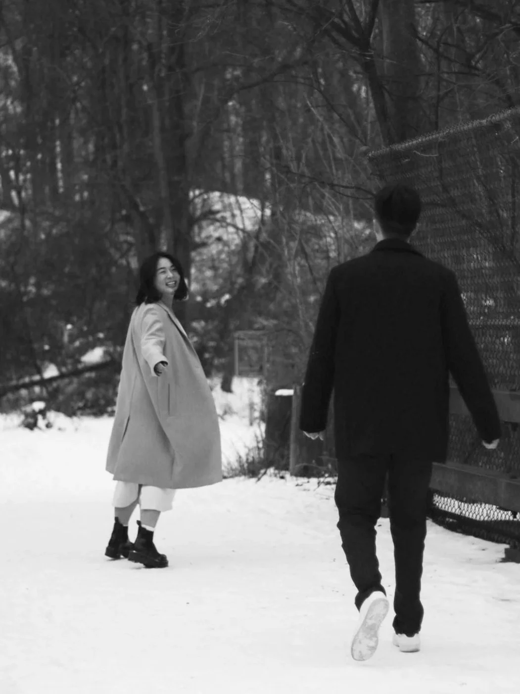 A black-and-white photo of a woman smiling and walking in the snow towards a man walking away from her. They are outdoors near a fence and trees.
