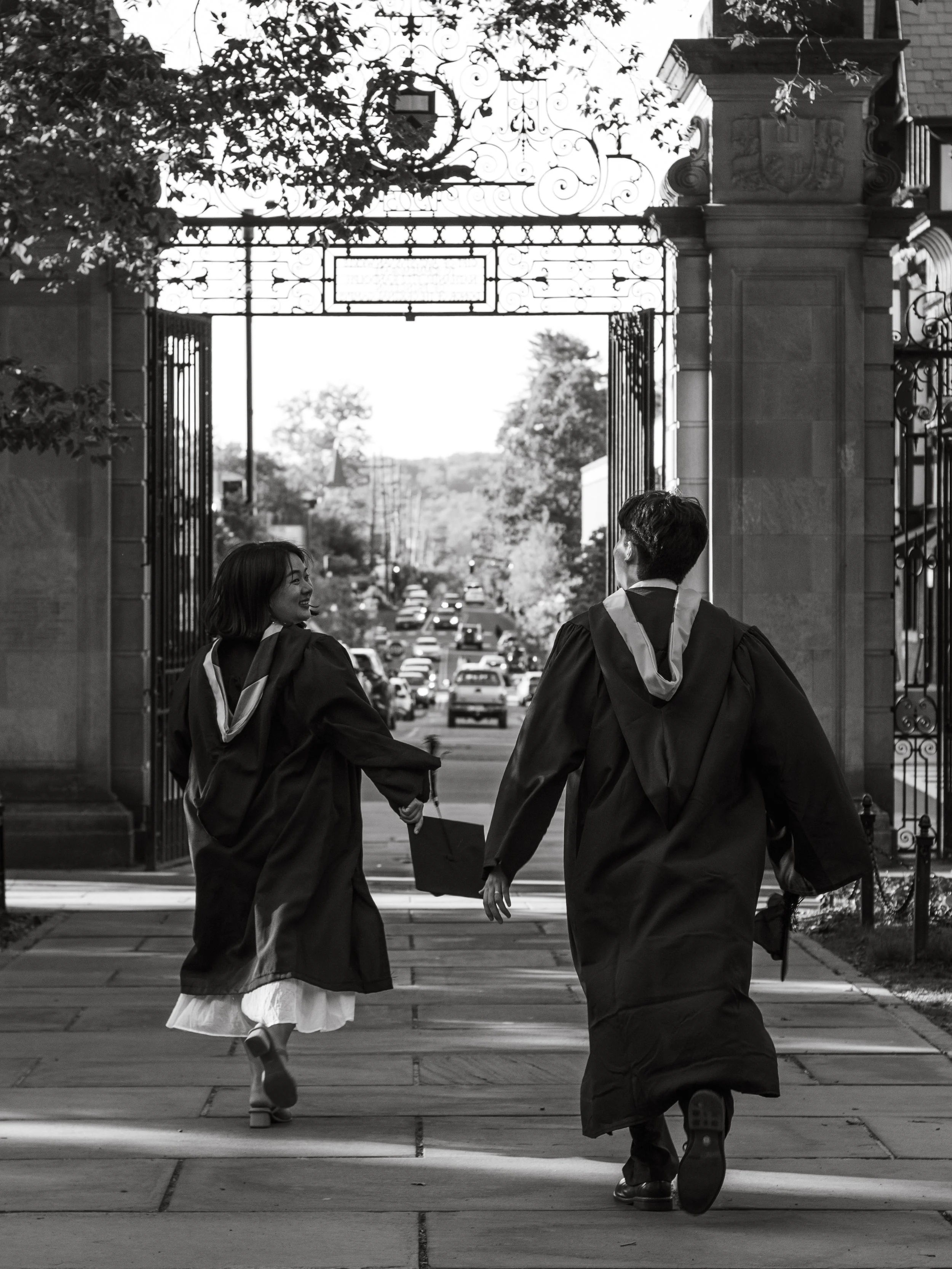 Two graduates in caps and gowns holding hands and walking together outdoors beneath an ornate gate, smiling and looking at each other.