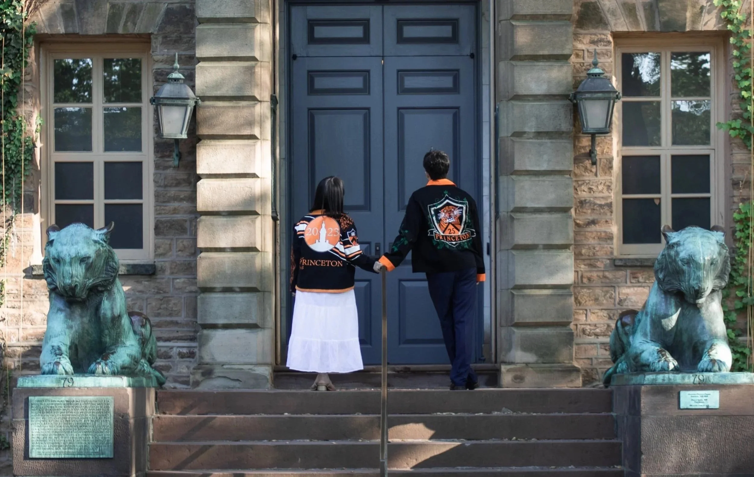 Two people standing in front of a large blue door on a brick building, holding hands. They are wearing Princeton jackets. Two lion statues are on either side of the stairs, and there are two lantern-style lights beside the door.