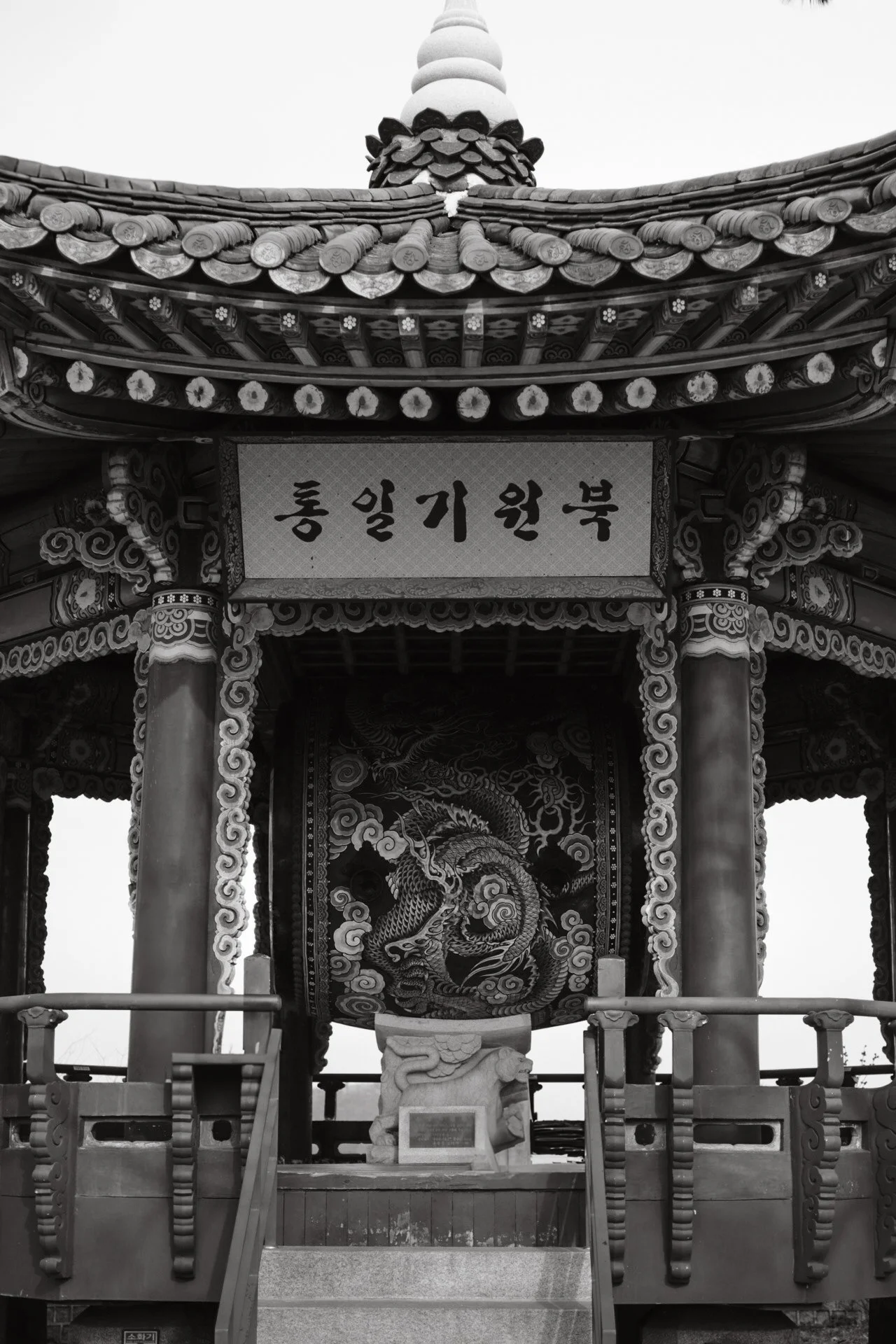Traditional Korean pavilion with intricate wooden carvings, supported by wooden columns, with a sign in Korean characters  and a detailed dragon artwork in the background.