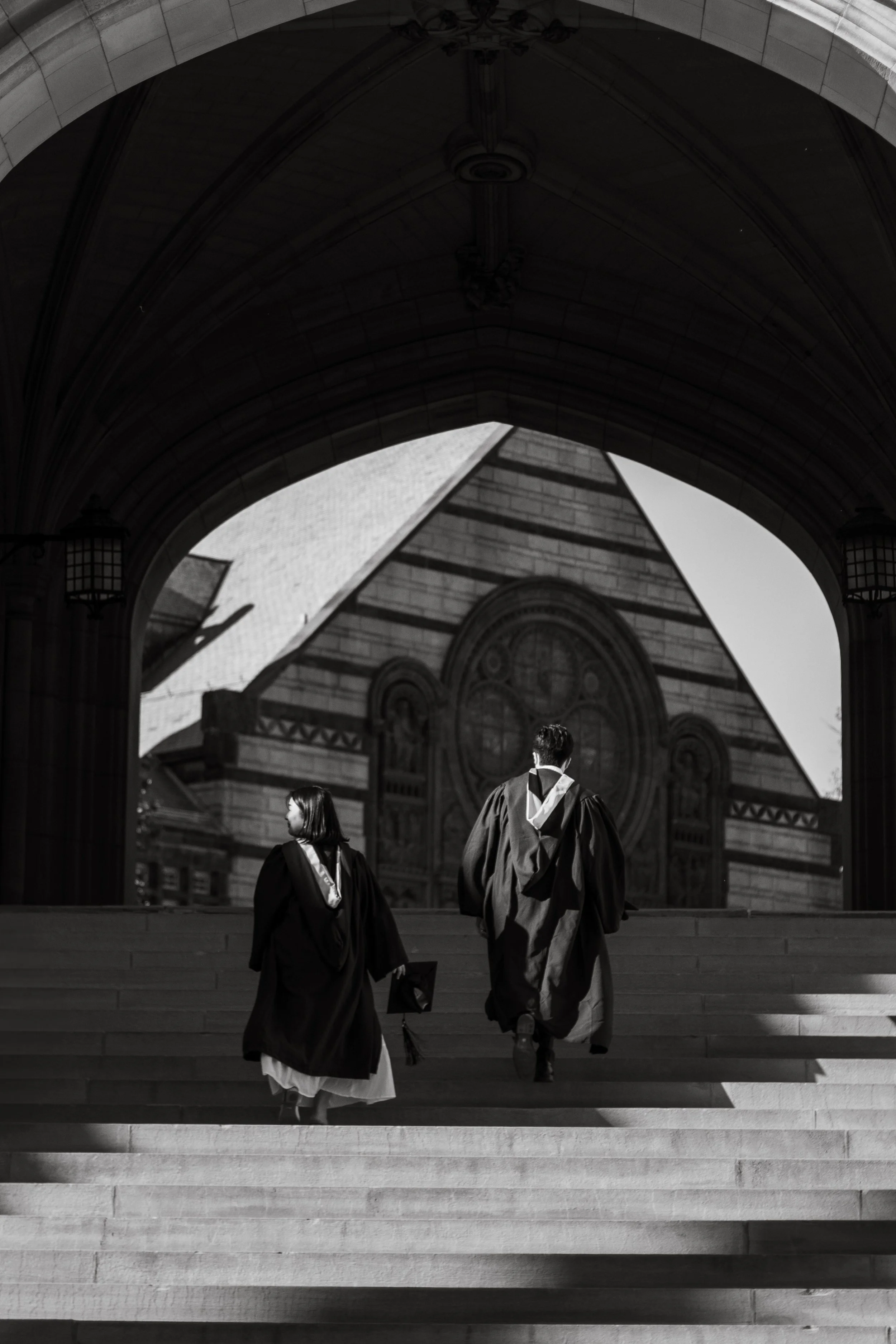 Black and white photo of three graduation graduates walking up stairs outside a historic building with arched doorway, ornate design, and clock on facade.