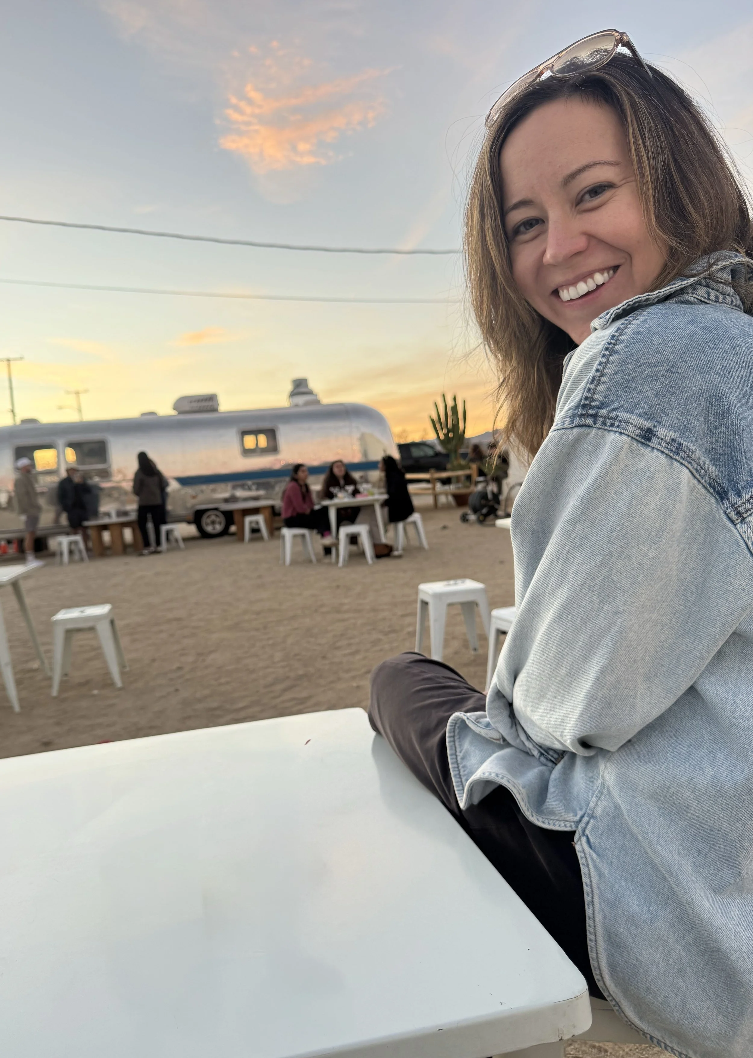 A smiling woman with brown hair, sunglasses on her head, wearing a light denim jacket, sitting outdoors at a white table during sunset. In the background, there are people sitting at tables, a vintage Airstream trailer, and a cactus, with a colorful sky.