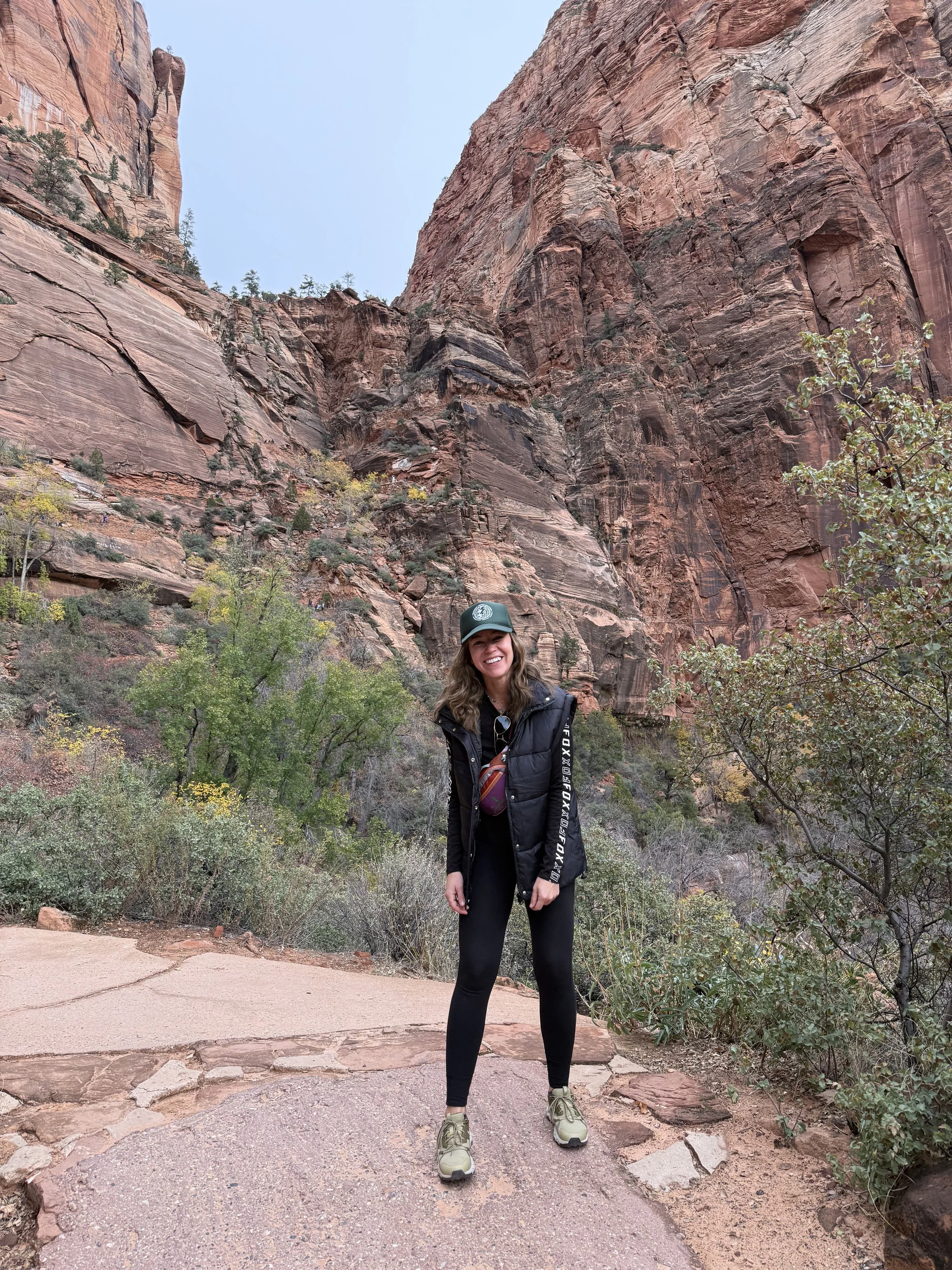 A woman smiling stands on a trail in a canyon with red rock cliffs and some green vegetation in the background.