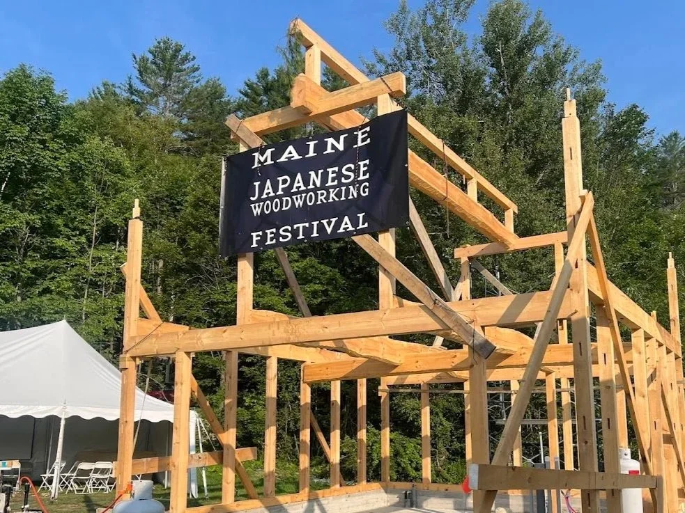 Wooden framework of a building under construction at the Maine Japanese Woodworking Festival, with a black banner hanging from the structure.