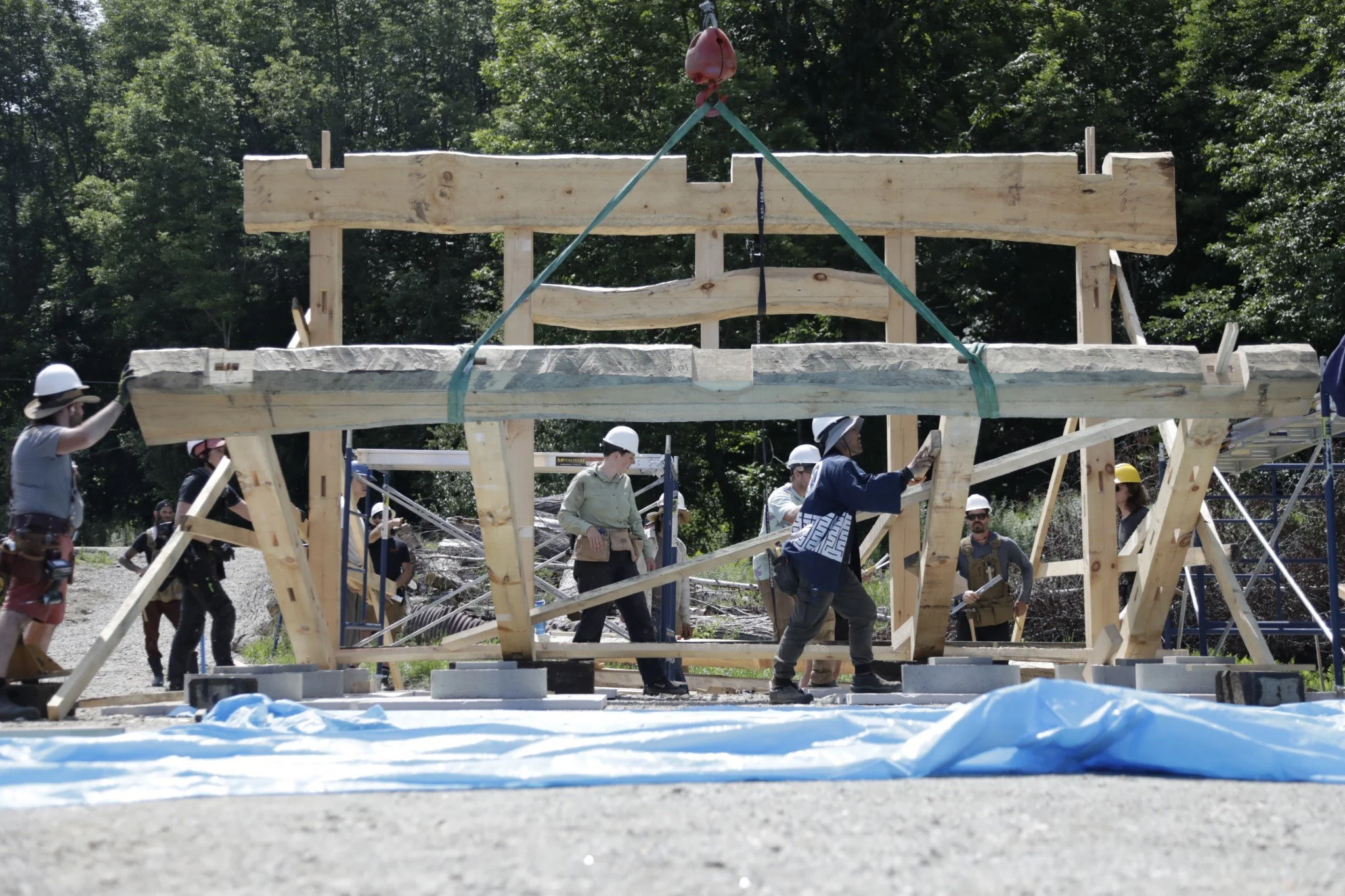 Construction workers wearing safety helmets and harnesses assembling a wooden structure outdoors with trees in the background.