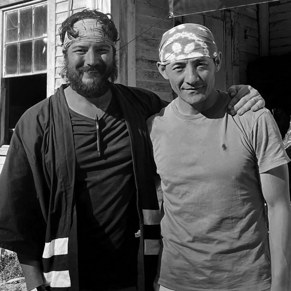 Two men standing close together outdoors, one with a beard and wearing a headband, the other with a bandana, both smiling, with a wooden building in the background.