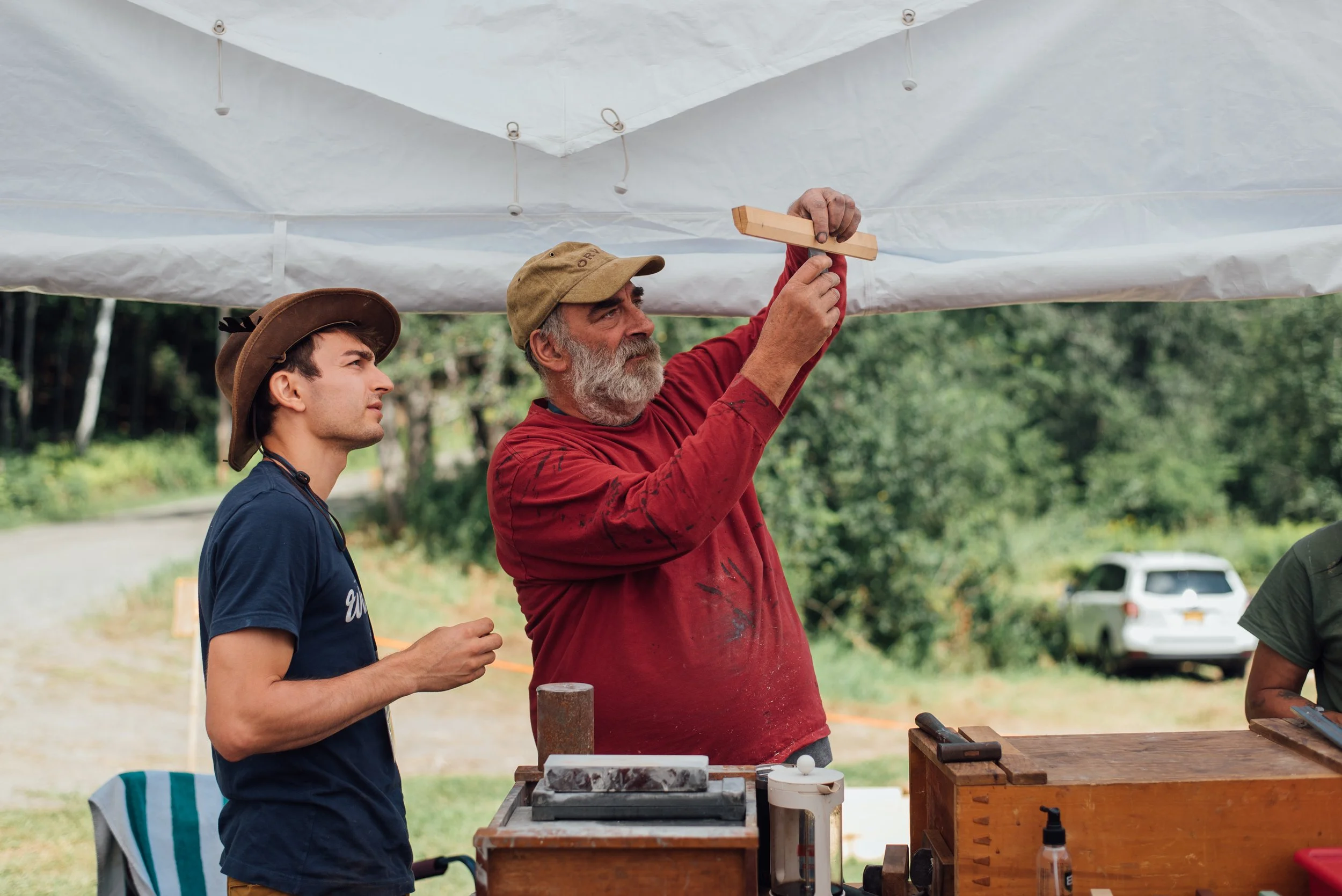 Two men, one with a beard and wearing a red shirt and another younger man in a navy shirt and a wide-brimmed hat, are working outdoors under a canopy. The older man is holding a wooden piece while the younger man observes attentively. There are tools and wood on a table in front.