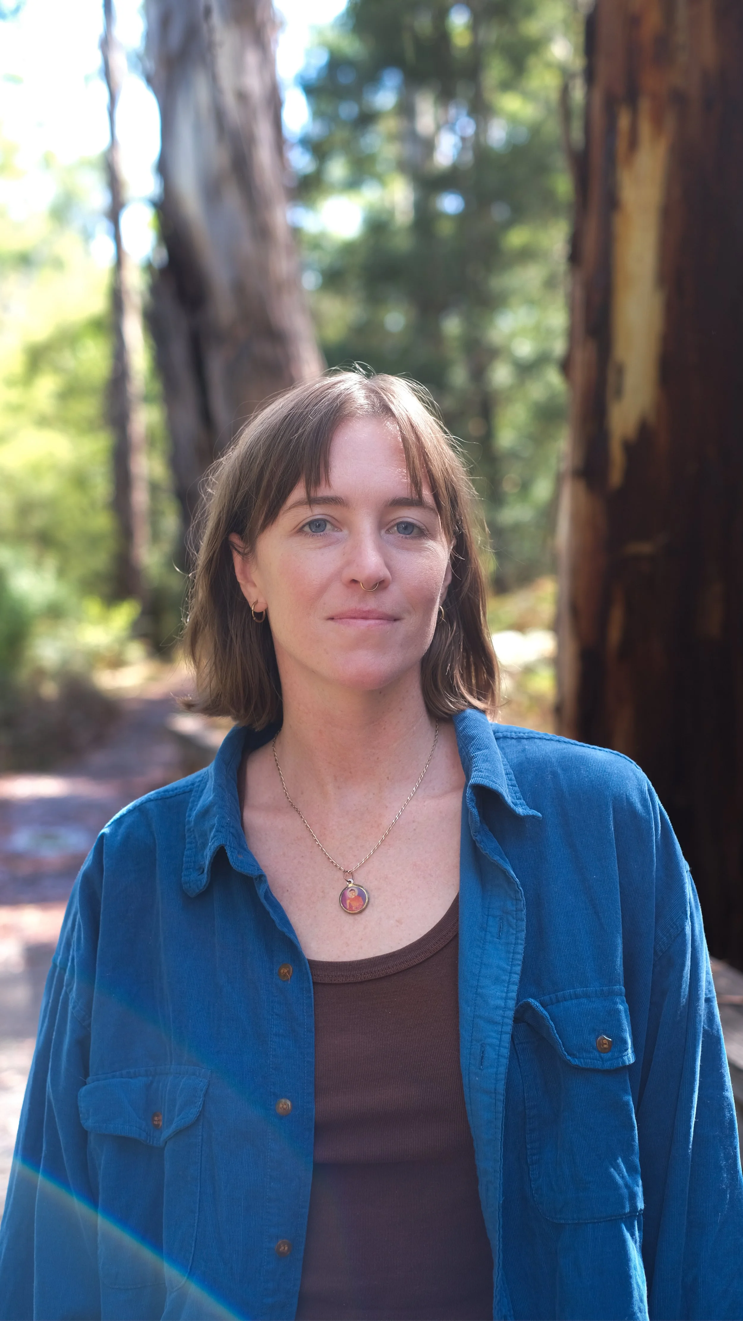 A woman with shoulder-length brown hair, light skin, and blue eyes standing outdoors in a forest near trees with thick trunks. She is wearing a blue button-up shirt over a brown top and has a pendant necklace and hoop earrings.