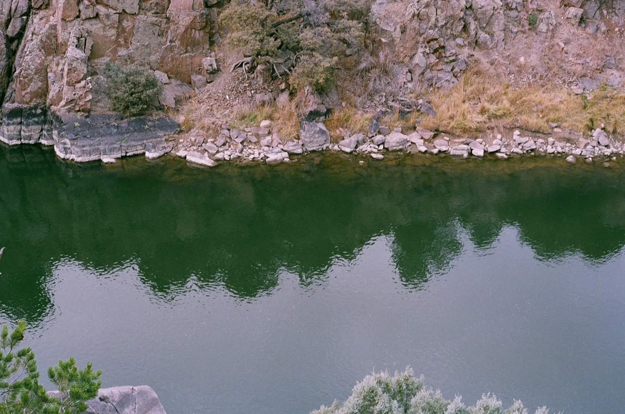 A body of water with a rocky, grassy shoreline and a cliff with trees reflected in the water.