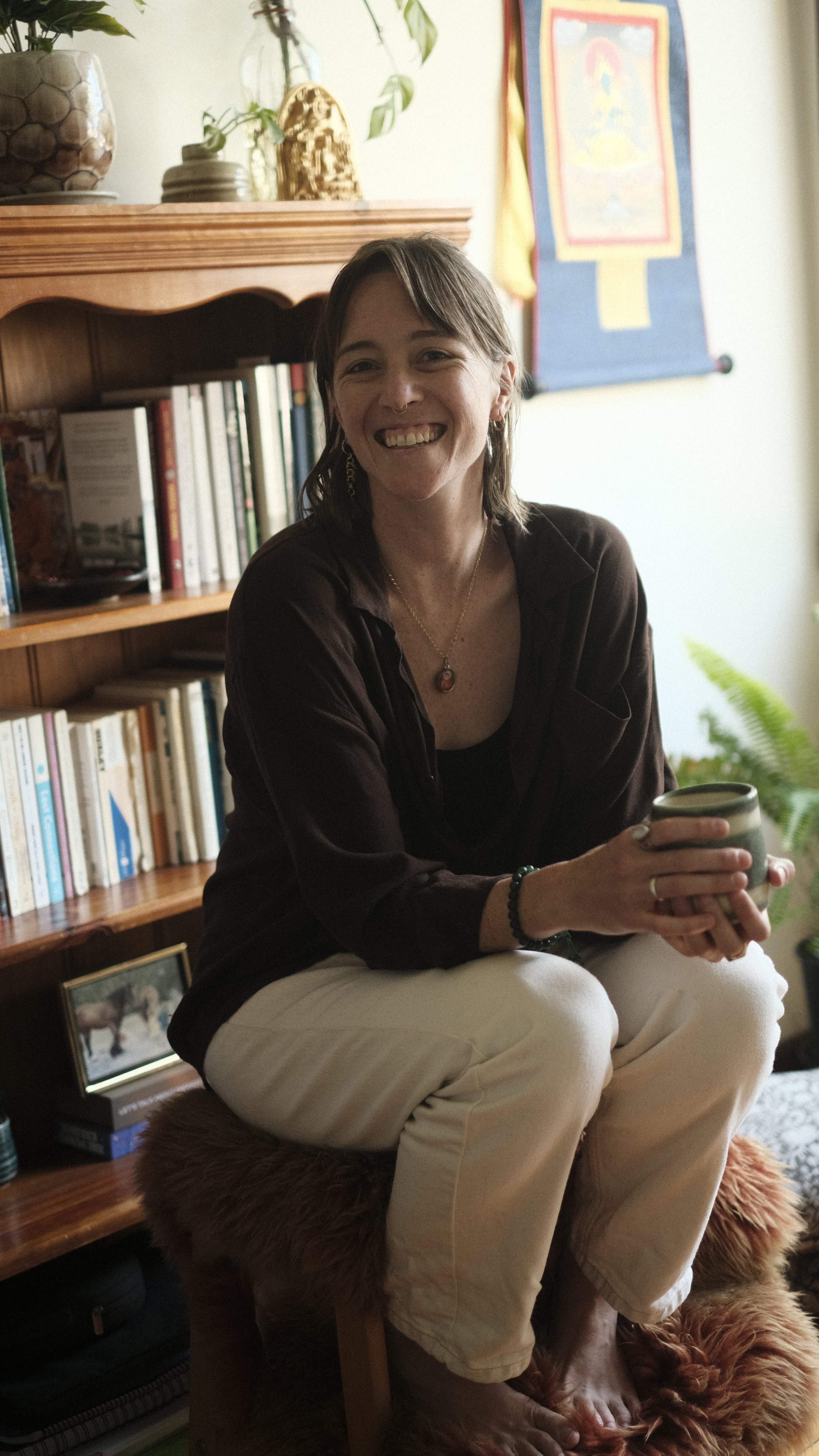 A smiling woman with shoulder-length brown hair, wearing a dark shirt and white pants, sitting on a sheepskin rug on a stool in a cozy room with bookshelves, plants, and a Thangka of Green Tara in the background, holding a mug.