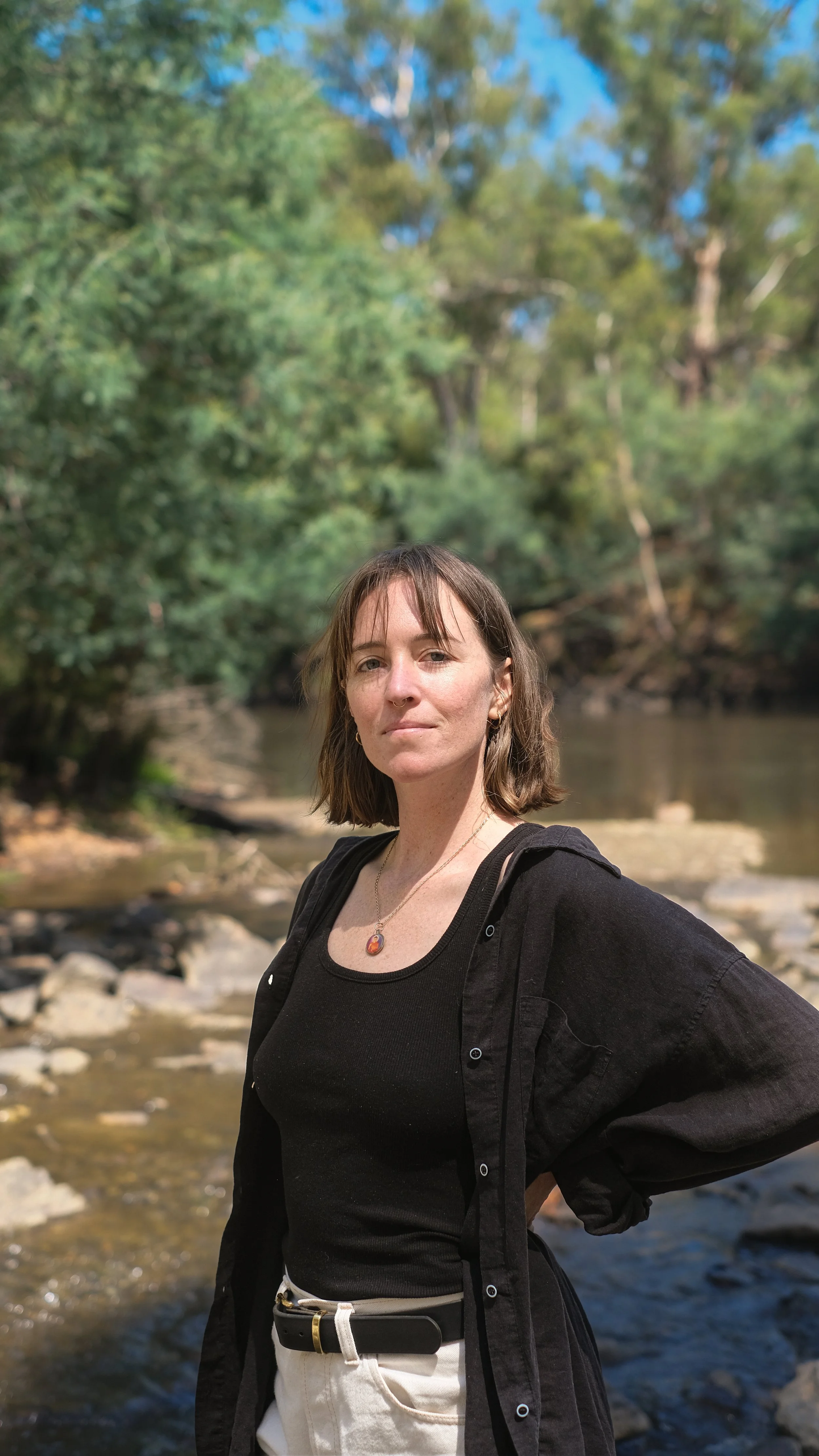 A woman with short brown hair standing by a river in a wooded area, wearing a black tank top, black shirt, white pants, and a necklace.