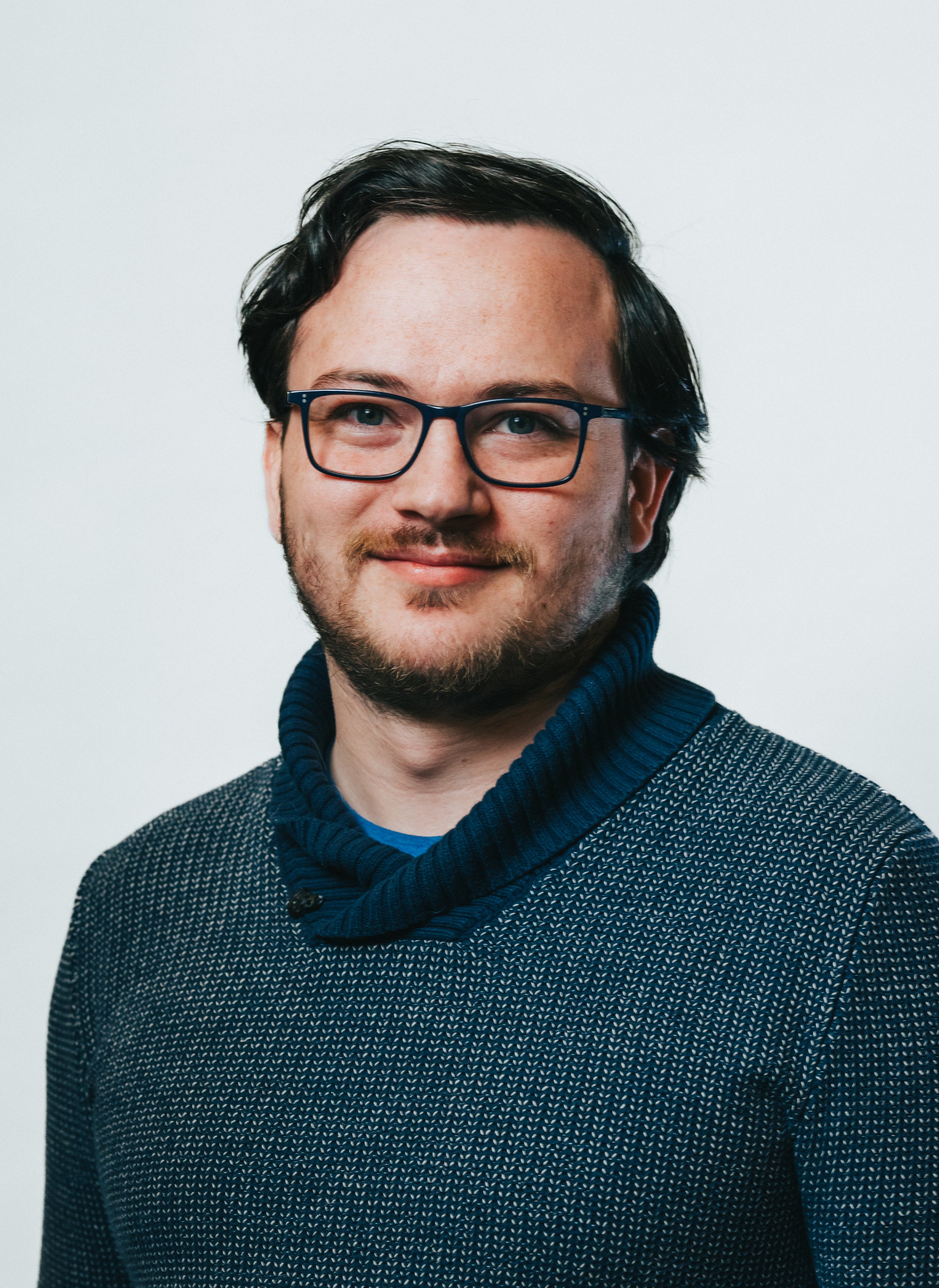 Zachary Sewell, a young man with glasses, dark hair, and a beard, wearing a dark blue patterned sweater with a high collar, smiling against a plain white background.