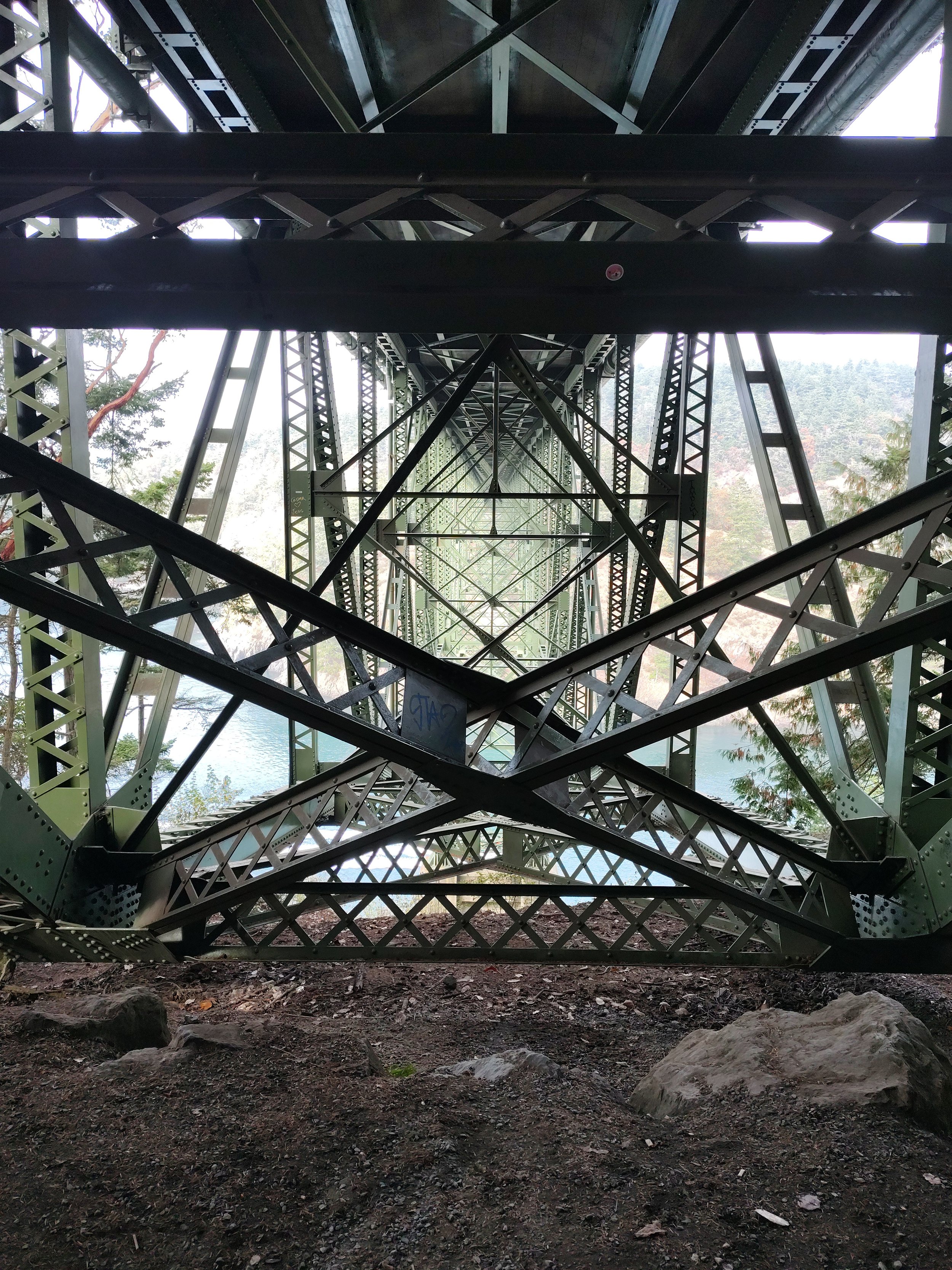 Underneath the Deception Pass bridge with visible trusses and support beams over a river, with trees and hills in the background.