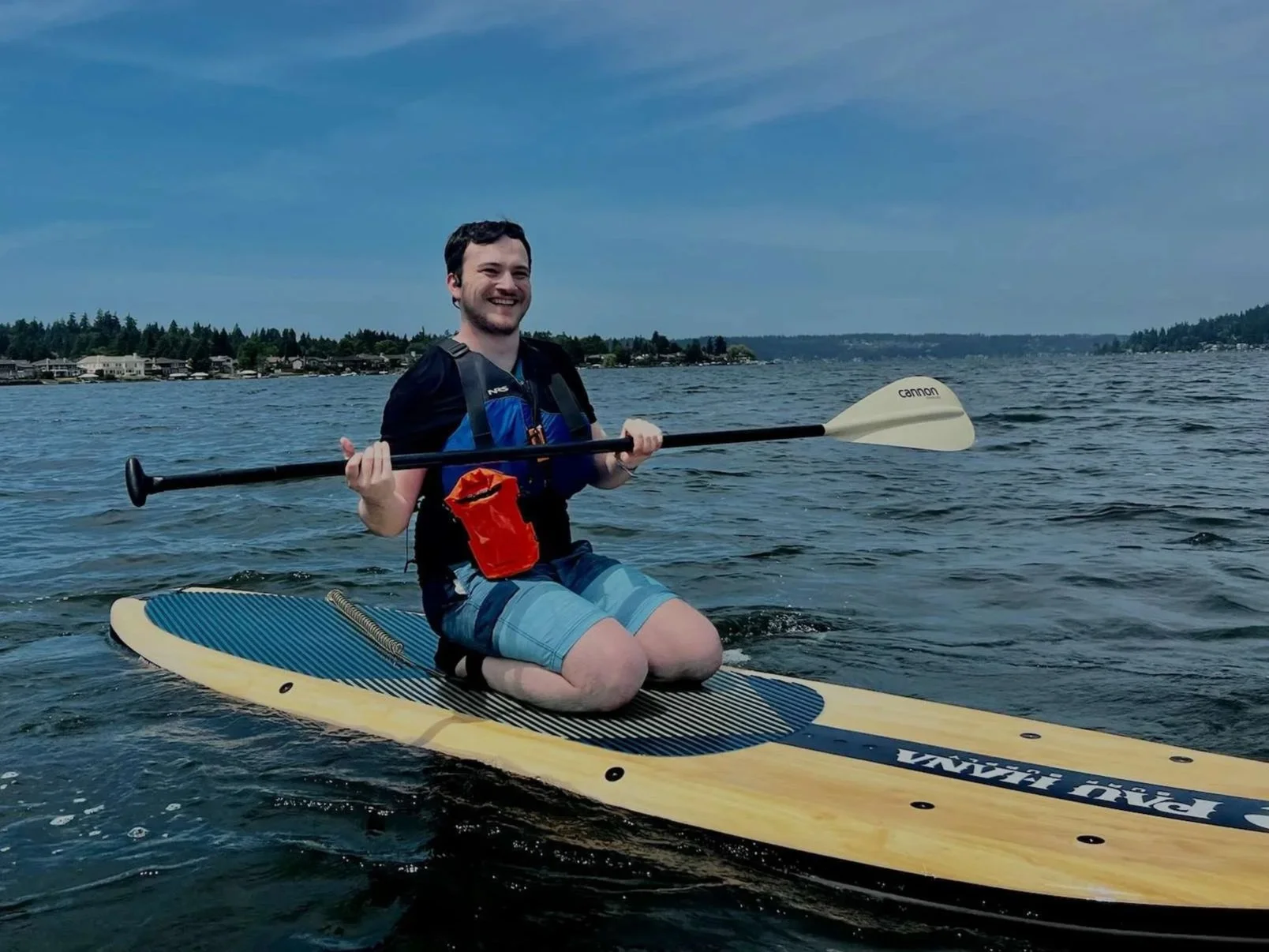 Zac on a paddleboard in a body of water, smiling and holding a paddle, with a shoreline and houses in the background.