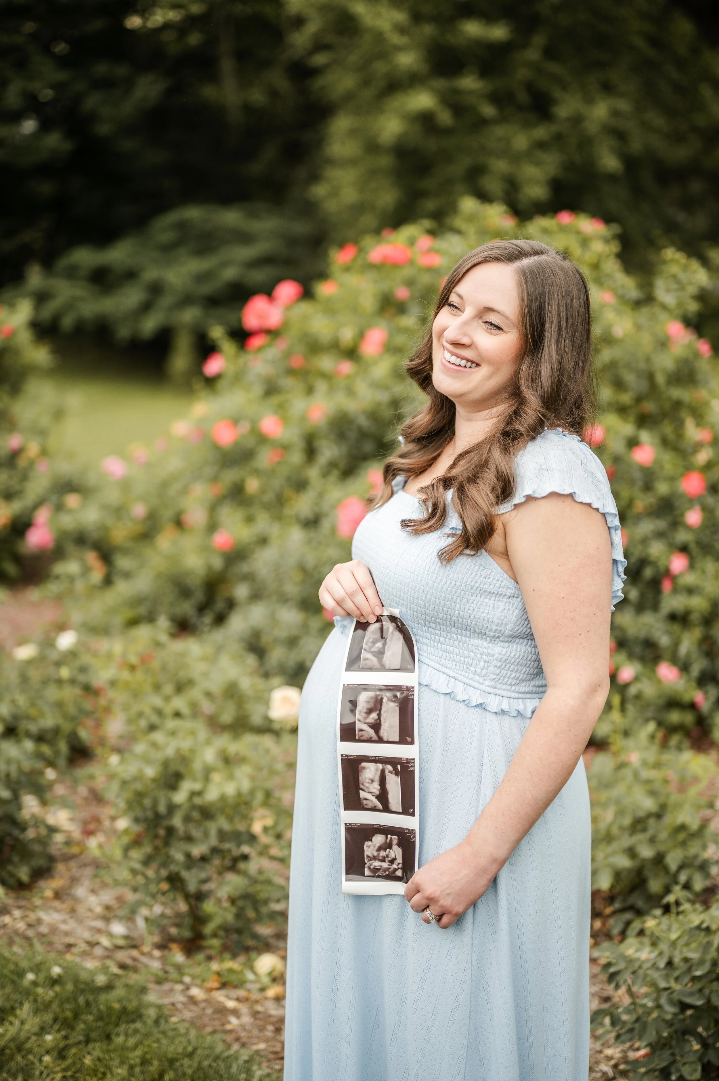 Pregnant woman smiling outdoors in a garden, holding ultrasound images against a backdrop of blooming roses and green foliage bon air rose garden arlington virginia NOVA
