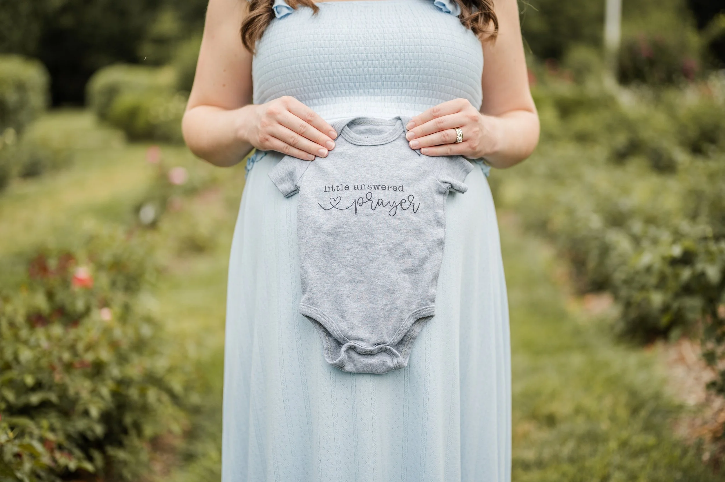 A pregnant woman in a light blue dress is holding a small gray baby onesie with the words "little answered prayer" embroidered on it, in an outdoor garden setting bon air rose garden arlington virginia NOVA