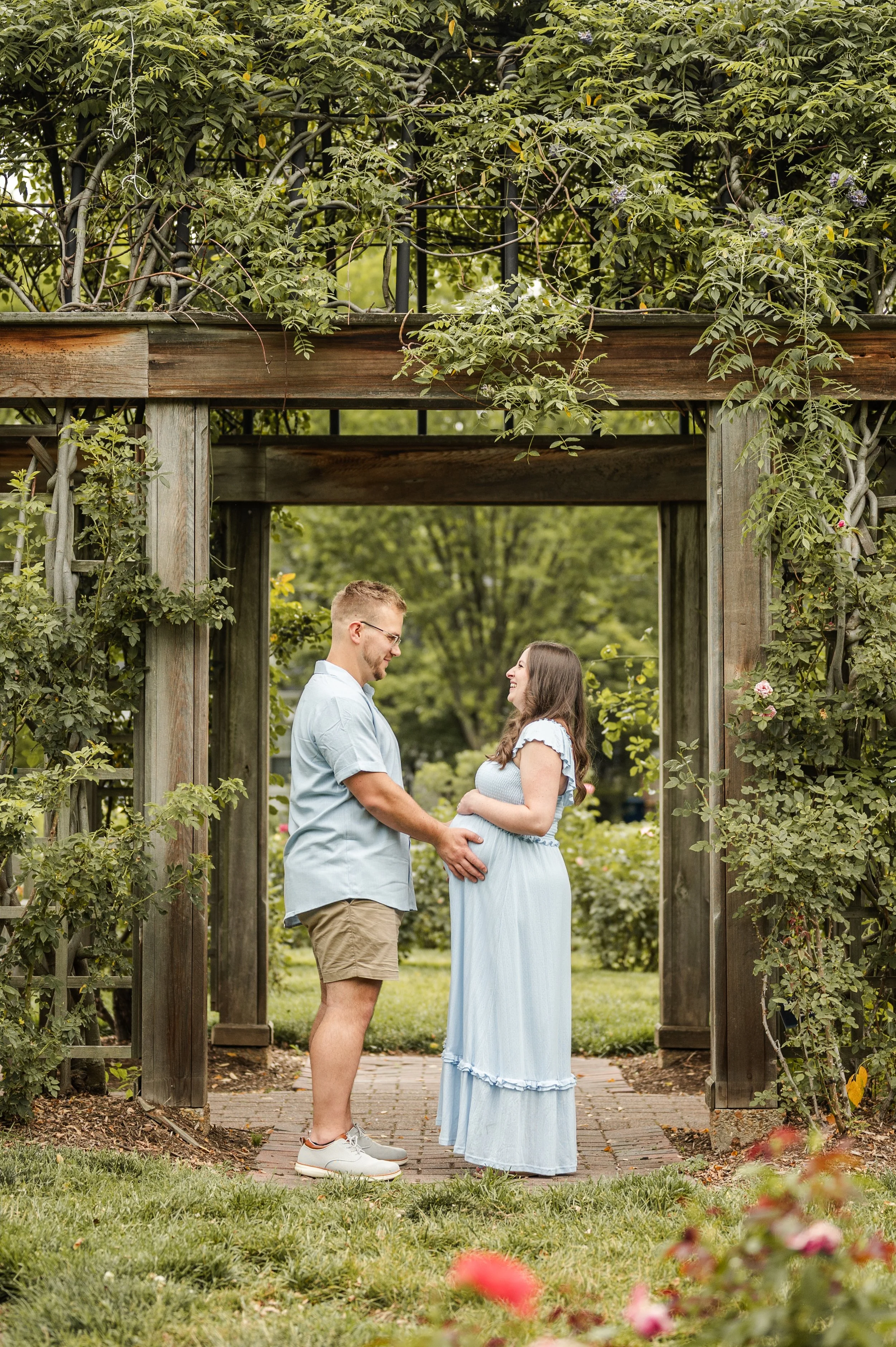 A couple stands face-to-face, holding hands and smiling at each other beneath a wooden archway covered with green vines and plants in a lush garden. The woman is pregnant, and they are both dressed casually, with the man wearing a light blue shirt an
