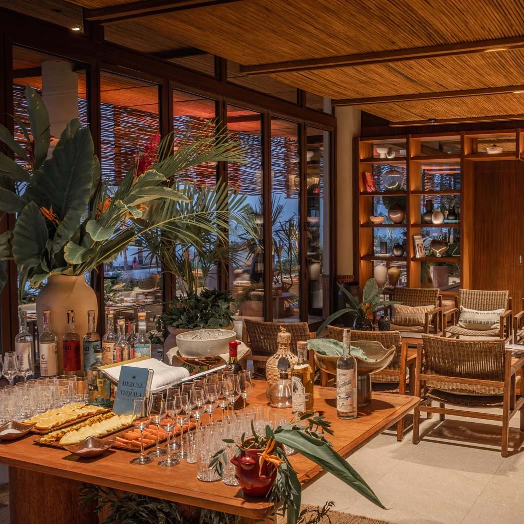 Interior of a stylish, cozy living room or lounge area with wooden furniture, large plants, and a bar setup with glasses and bottles, featuring warm lighting and large windows with venetian blinds.