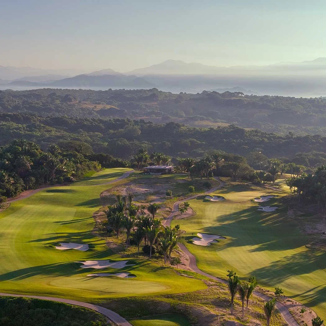 Aerial view of a golf course with sand bunkers, palm trees, and a clubhouse, surrounded by lush greenery and distant mountains.