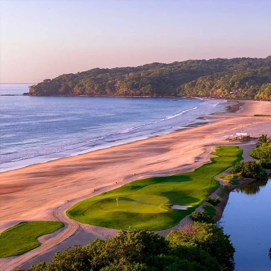 A scenic view of a coastal golf course with lush green fairways and a sand bunker, adjacent to a sandy beach and ocean, with a forested hill in the background at sunset.