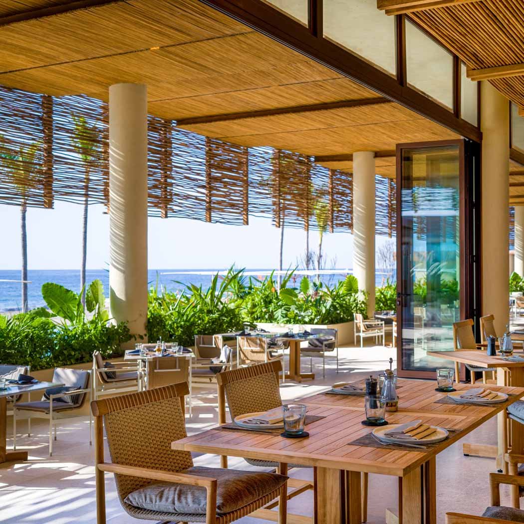 Interior of a seaside restaurant with wooden furniture, large glass doors, lush greenery, and an ocean view with palm trees in the background.