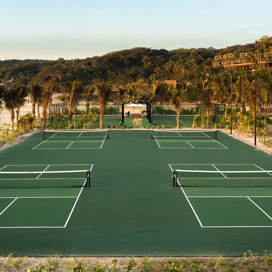 Four tennis courts with nets, surrounded by palm trees and greenery, with a hillside and some houses in the background during sunset.