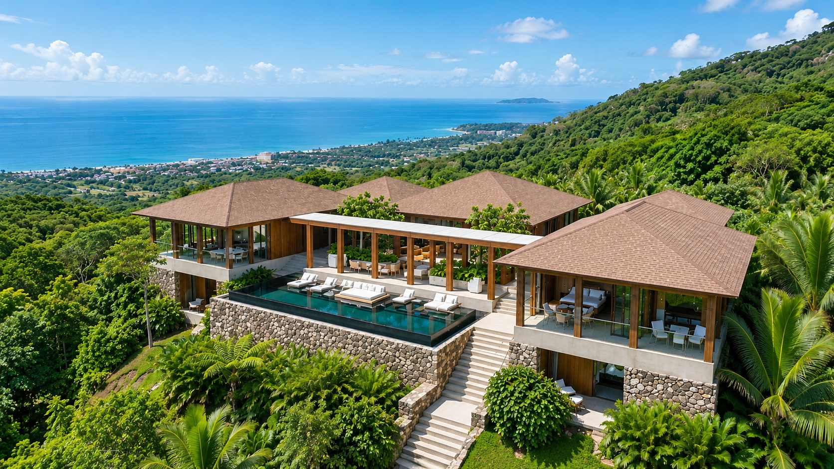 A luxurious hillside house with multiple levels, large deck with outdoor furniture, infinity pool, surrounded by lush greenery, overlooking the ocean and distant shoreline under a partly cloudy sky.