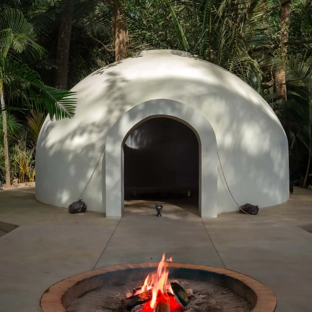 A white, dome-shaped structure with an arched entrance, situated outdoors in a lush tropical environment. In front of the structure, there is a fire pit with a small fire burning, and the structure is secured with ropes and weights.