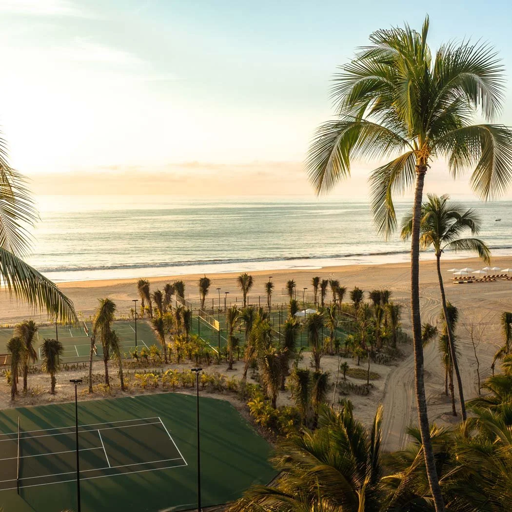 A tropical beach with tall palm trees, tennis courts, and lounge chairs near the ocean at sunset.