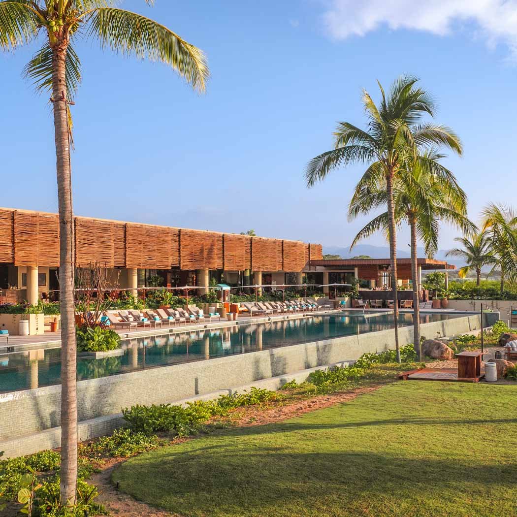 Luxury resort with a swimming pool, palm trees, lounge chairs, and a building with a thatched roof, set against a clear blue sky.